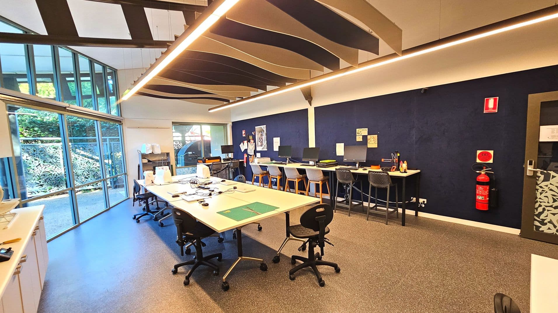 A modern workspace with a high ceiling featuring wave-like acoustic panels. The room has a large glass wall on the left, allowing natural light to illuminate the space. Several workstations with computers and chairs line the dark blue wall on the right. A central table with chairs is in the foreground. The floor is covered with speckled grey tiles, and a fire extinguisher is mounted on the wall near a patterned glass door.