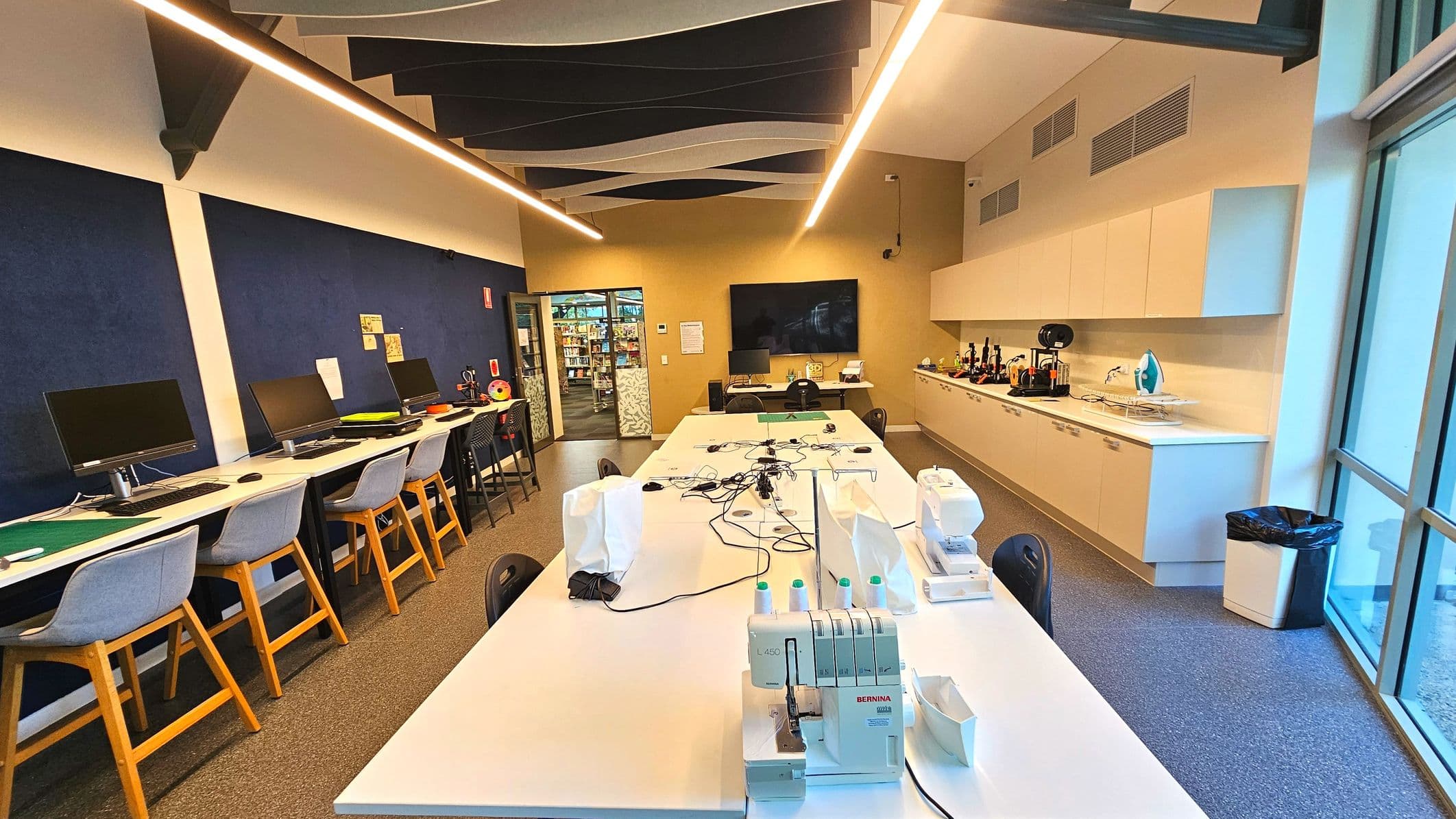 A modern workspace with a long central table equipped with sewing machines and cables. On the left, a row of computers with grey chairs is set against a dark blue wall. The right side features a countertop with 3D printers and an iron, beneath white cabinets. The room has a high ceiling with wave-like panels and is enclosed by glass walls on one side. A doorway at the back leads to a library area.