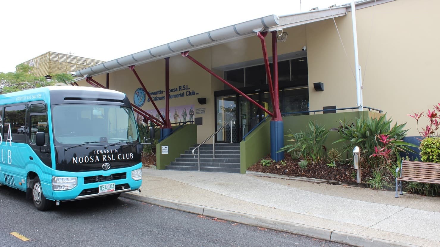 Another view of the previous image focuses on the glass sliding door and black tiled steps under the red/blue pillar-supported exterior. A steel rail in the centre divides the stepway into two halves bordered by green-painted half-walls with rails on both sides. Plants are in triangle forms by a seat on the right facing the sidewalk and parking lot.  The bus is packed in front.