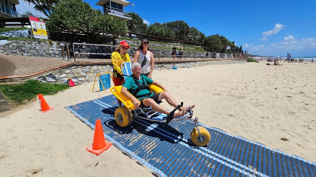 A man is seated in a beach wheelchair with large yellow wheels on a blue mat laid over the sand. Two women stand behind him, one wearing a lifeguard uniform. Orange cones mark the path. In the background, a stone wall with a railing separates the beach from a grassy area. A sign with beach safety information is visible, and there are people and beachgoers in the distance near the water.