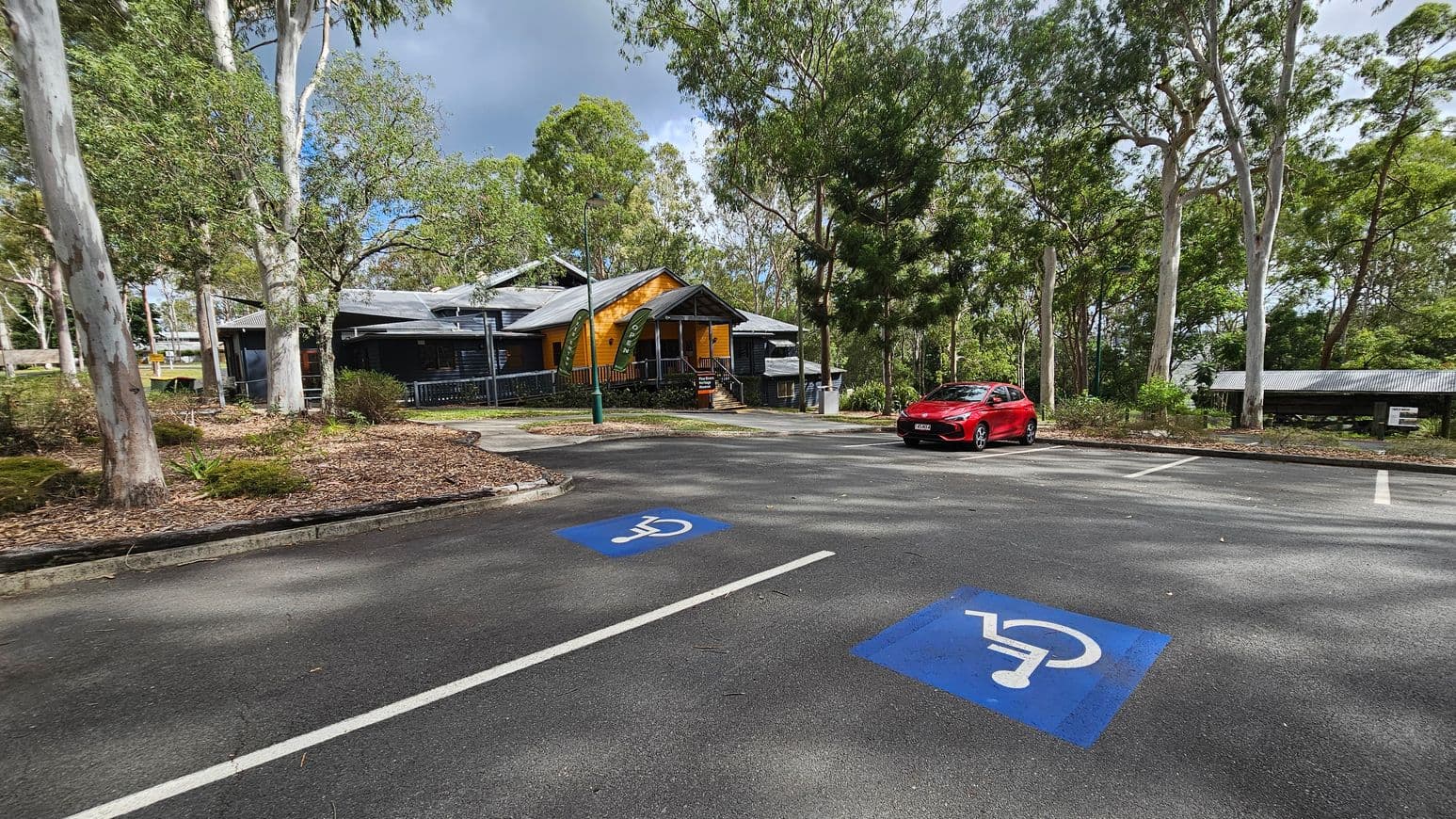 A building with a bright yellow facade and a grey roof, featuring a central entrance with steps leading up to it. On the left, two green flags display the words "FREE ENTRY" and "OPEN". A sign in front reads "Pine Rivers Heritage Museum" with "Moreton Bay" below. The surrounding area has trees and bushes, with a path leading to the entrance. The building has a veranda with grey railings. The ground is covered with mulch and grass.