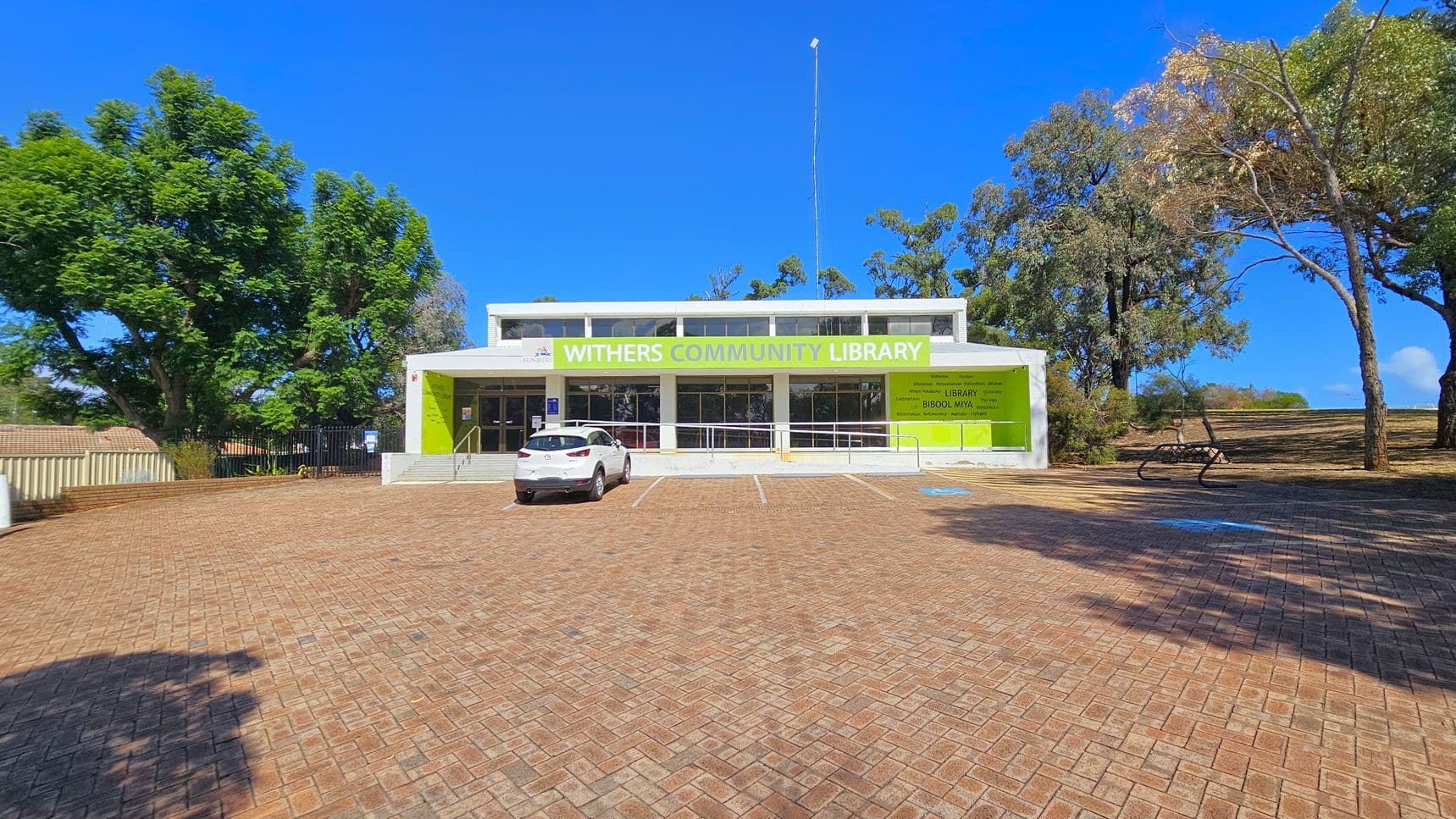 A paved pathway leads to a building in the centre, partially obscured by trees. On the left, a white car is parked near a beige fence, surrounded by trees. On the right, a green bin is placed on a concrete path, with more trees and a grassy area. Two blue signs are visible near the building, which has a sign reading "COMMUNITY" above the entrance. The ground is covered with fallen leaves and the sky is clear.