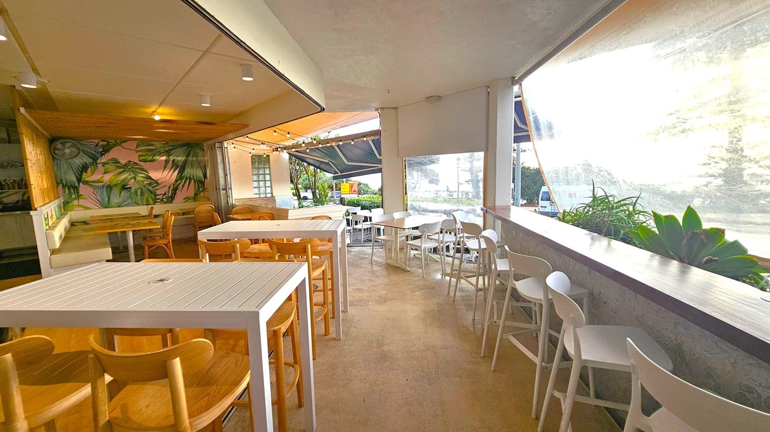 A café interior with a tropical theme. On the left, there's a seating area with white chairs and a wooden bench against a wall with neon signage. The middle features wooden tables and chairs on a wooden floor, with a wall decorated with large leaf patterns. A neon sign with palm tree imagery is on the back wall. On the right, a folding door is partially open. The foreground has a beige carpeted floor and a signpost. A dog is partially visible at the bottom.