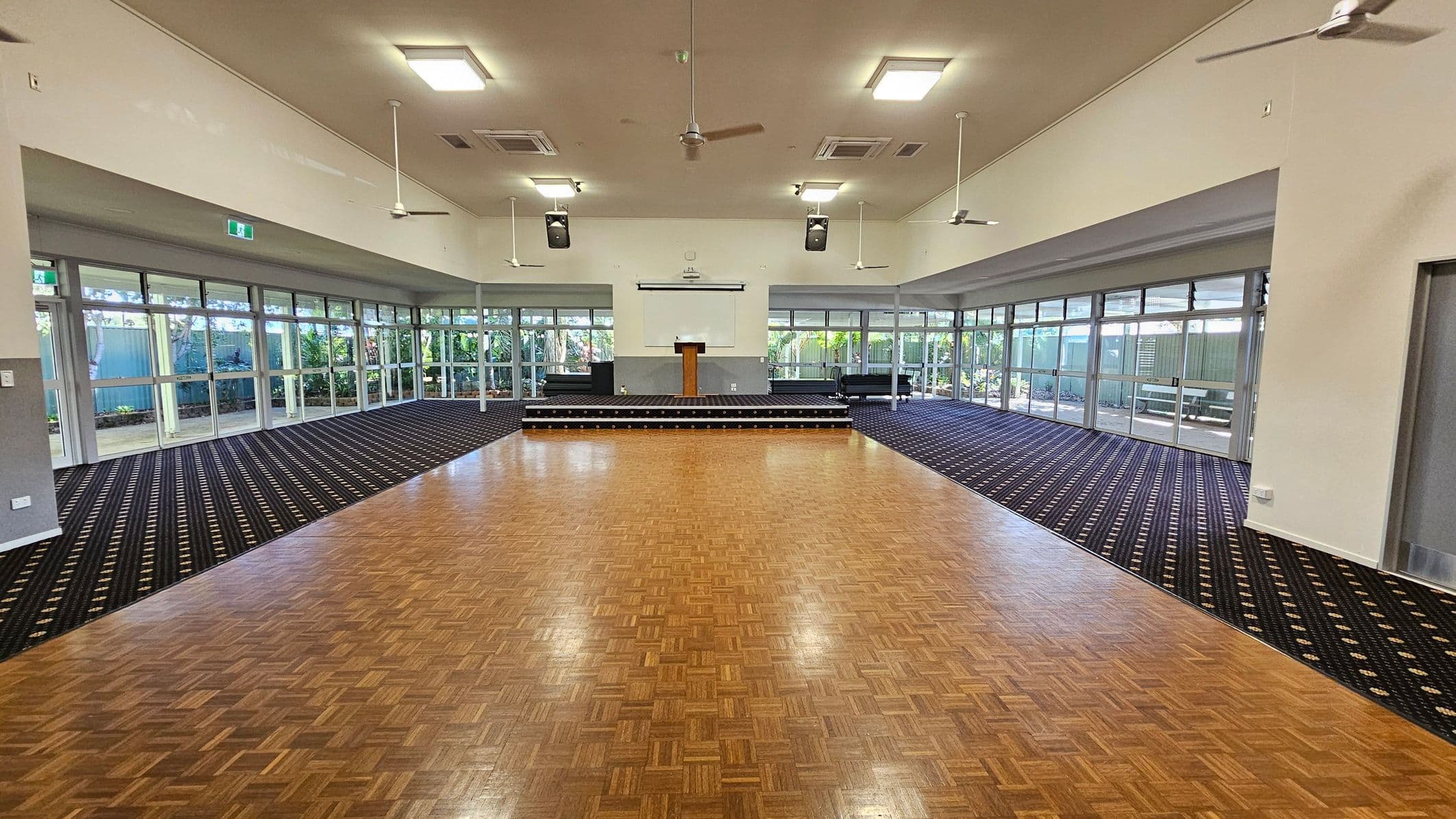 A spacious hall with a wooden parquet floor in the centre and patterned carpet on the sides. At the far end, there's a small stage with a lectern. Large windows line the left and right walls, allowing natural light to enter. Ceiling fans and lights are evenly spaced across the ceiling. The walls are light-coloured, and there are exit signs above the doors. The background shows greenery outside the windows.