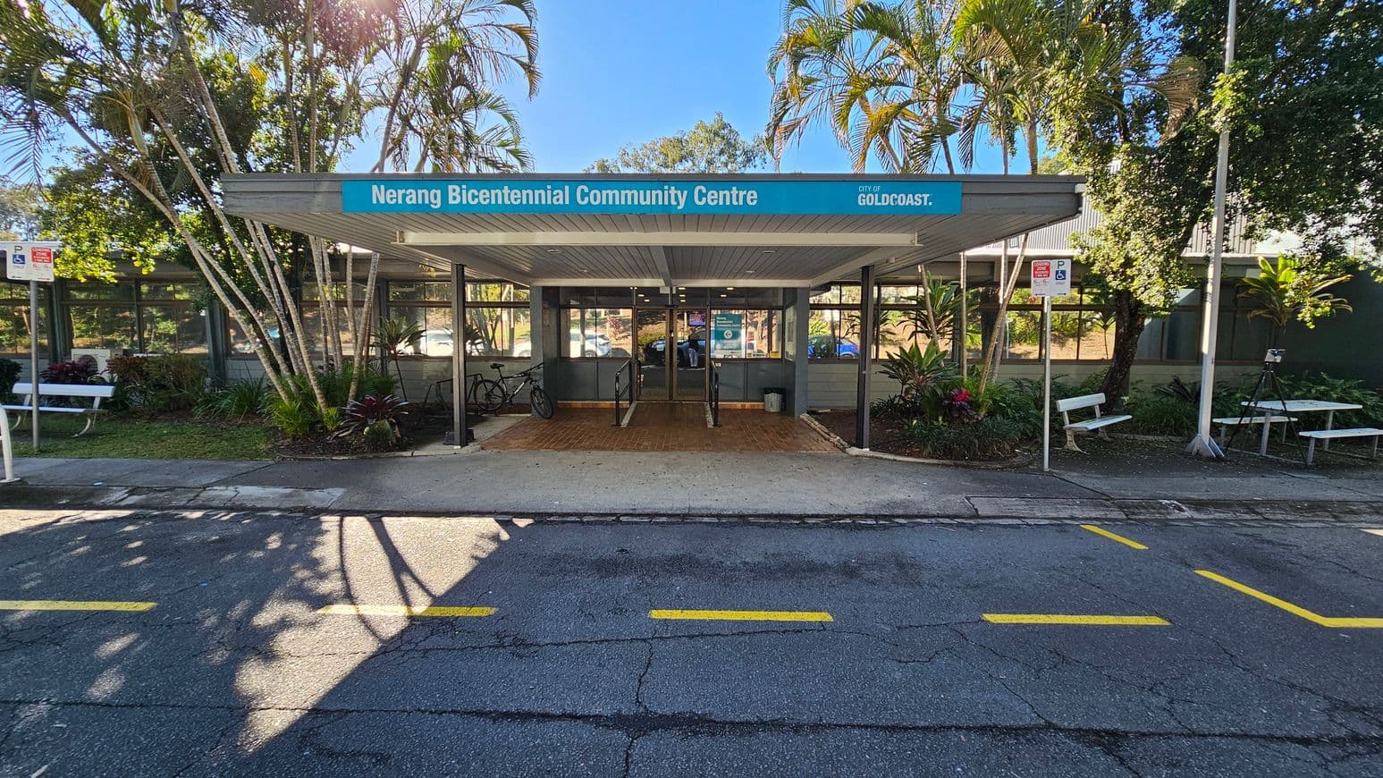 Entrance to the Nerang Bicentennial Community Centre with a blue sign above. The building has large windows and a covered walkway leading to the entrance. Bicycles are parked on the left. Palm trees and other greenery surround the area. Two white benches are on either side, with a picnic table on the right. The foreground shows a road with yellow markings and shadows from the trees.