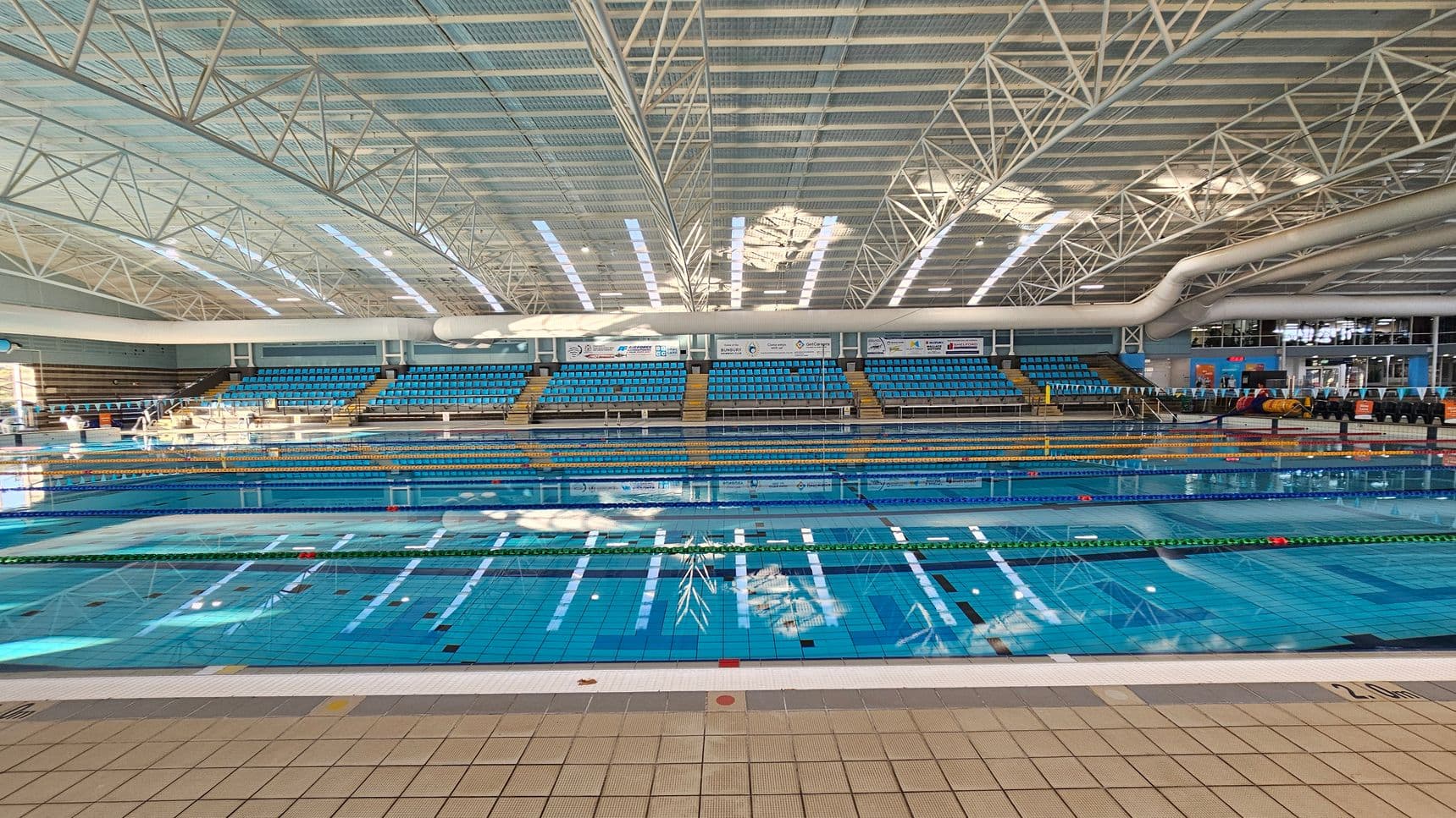 An indoor swimming pool with clear blue water, divided into lanes by floating lane markers. The background features tiered seating with blue chairs, and a large scoreboard or display area above. The ceiling is high with a grid of metal beams and skylights allowing natural light to filter through. The floor around the pool is tiled in a light colour.