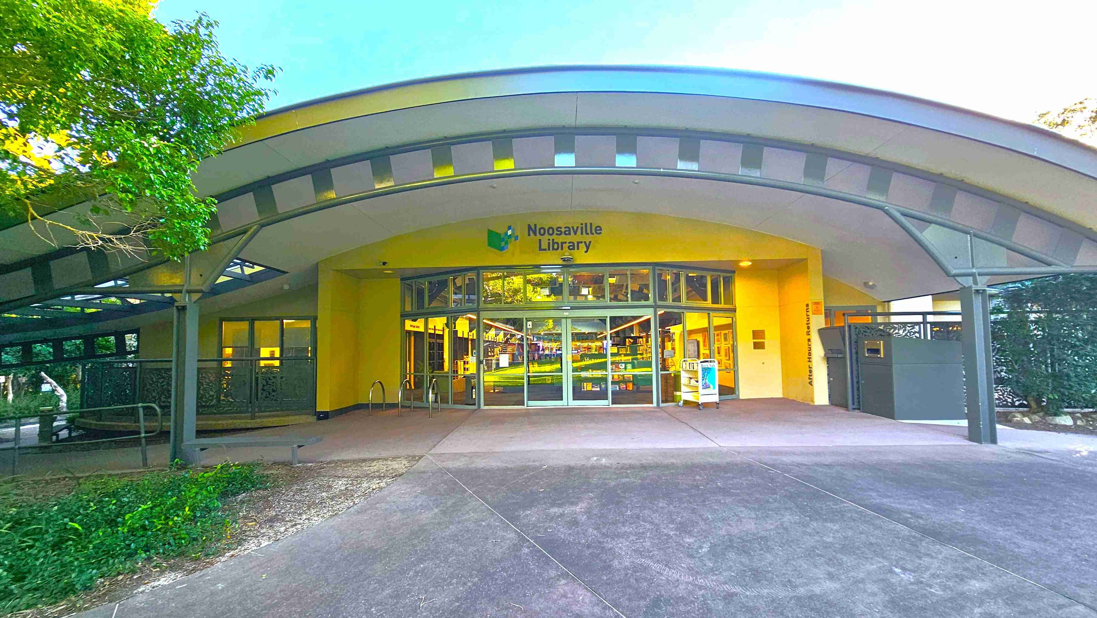 The entrance to Noosaville Library features a wide, curved metal awning with a geometric pattern, providing shade over the glass double doors. The library's name is displayed above the entrance. To the right, there's an after-hours book return slot. A bicycle rack is visible on the left, and the ground is paved with concrete. The area is bordered by greenery, creating a semi-enclosed feel.