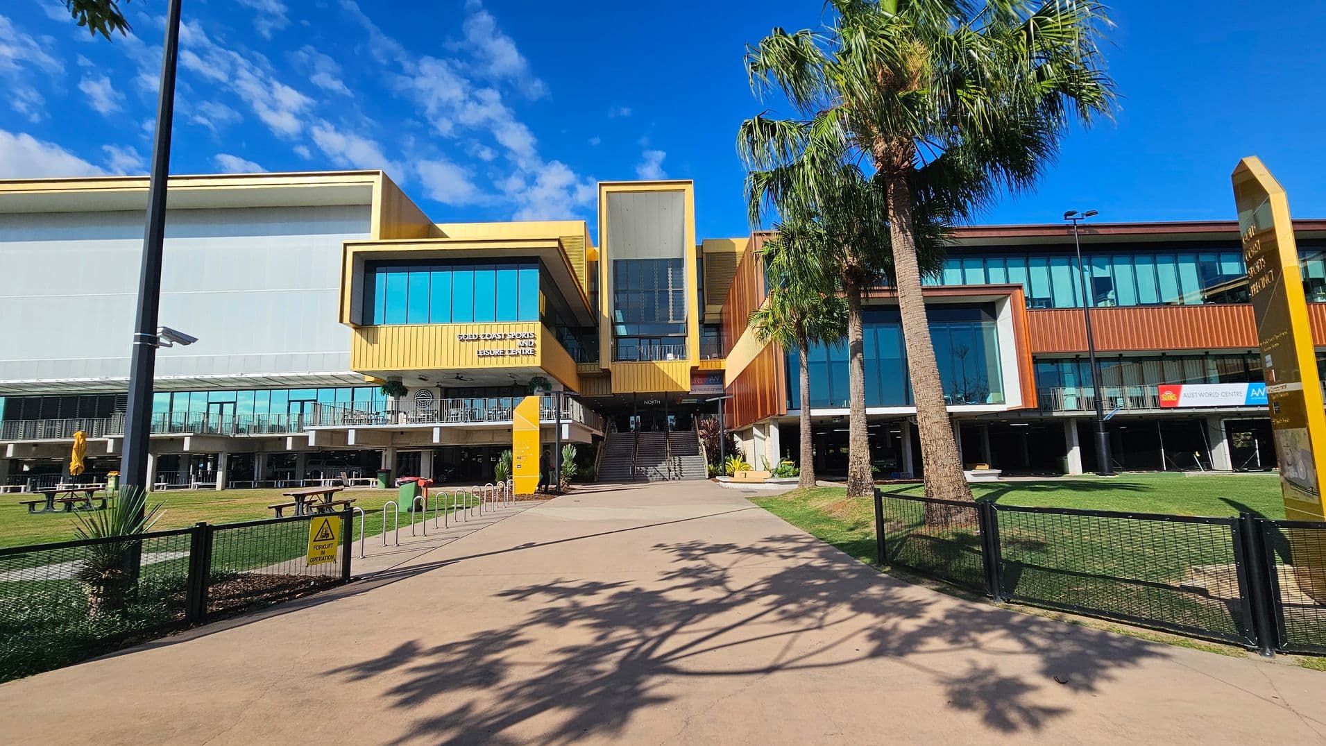 A modern building with a yellow and brown facade, featuring large glass windows. In the centre, a staircase leads up to the entrance. On the left, there is a grassy area with picnic tables and a yellow umbrella. On the right, palm trees and a yellow sign are visible. The foreground has a paved walkway with shadows of palm trees. A black fence lines the path.