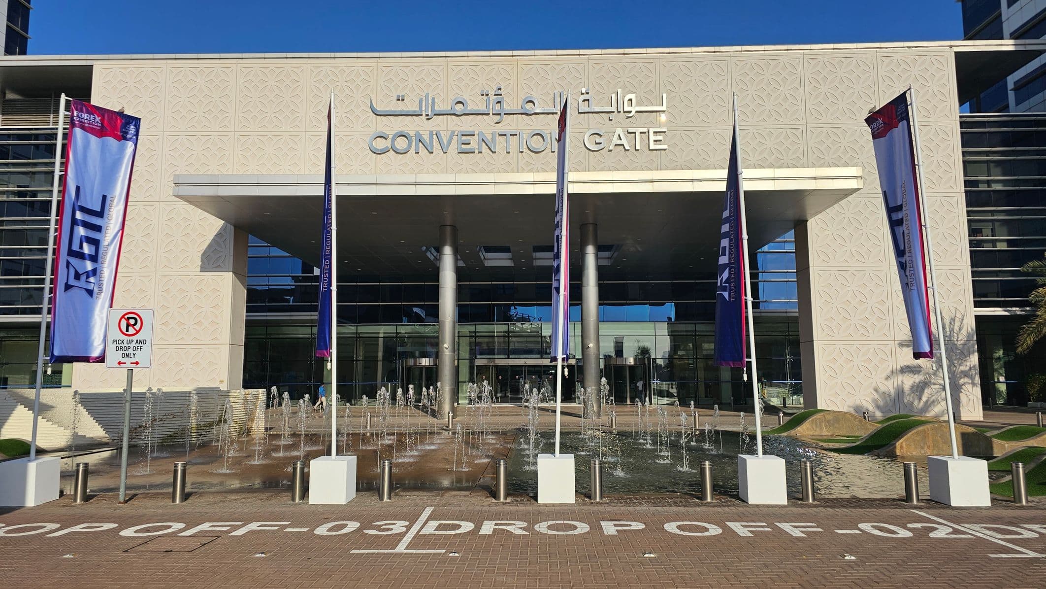 The image shows the entrance to a building labelled "CONVENTION GATE" with Arabic text above. The facade is light-coloured with a geometric pattern. In front, there are several vertical flags with text, a no parking sign, and a series of small fountains. The ground is paved with marked drop-off zones. The entrance has large glass doors and is supported by silver columns. On the right, there are some palm trees visible.