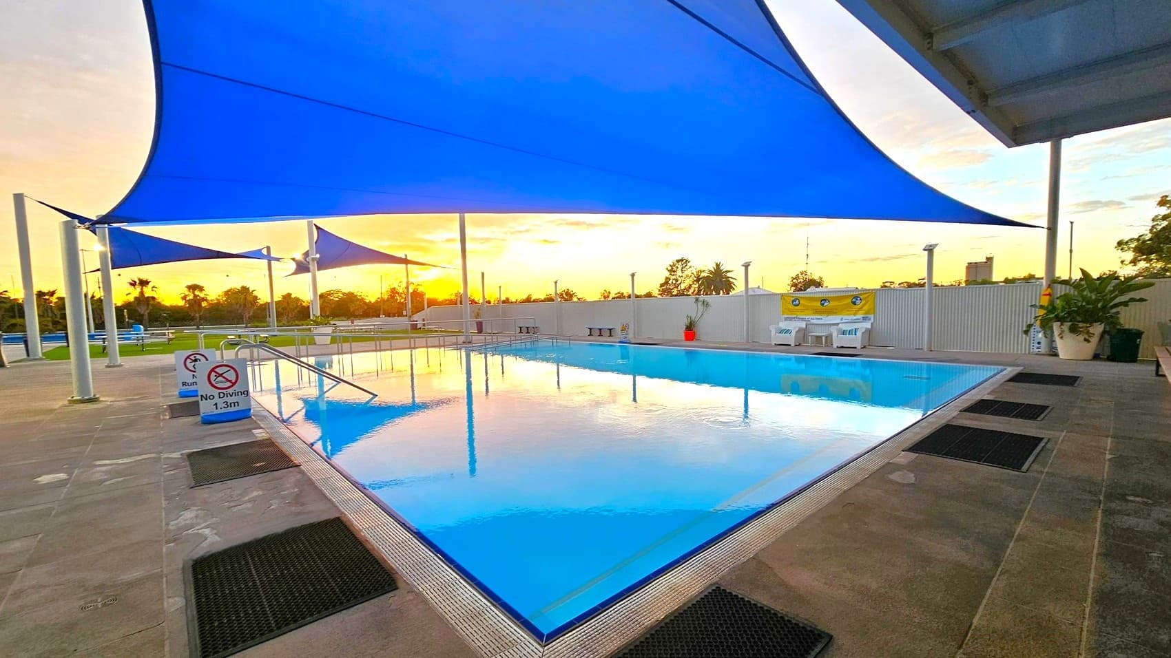 A swimming pool with clear water is in the centre, surrounded by a tiled deck. Blue shade sails are stretched above, providing cover. On the left, signs indicate "No Diving" and "No Running." In the background, a fence with a yellow banner and some potted plants are visible. The sun is setting, casting a warm glow over the scene.