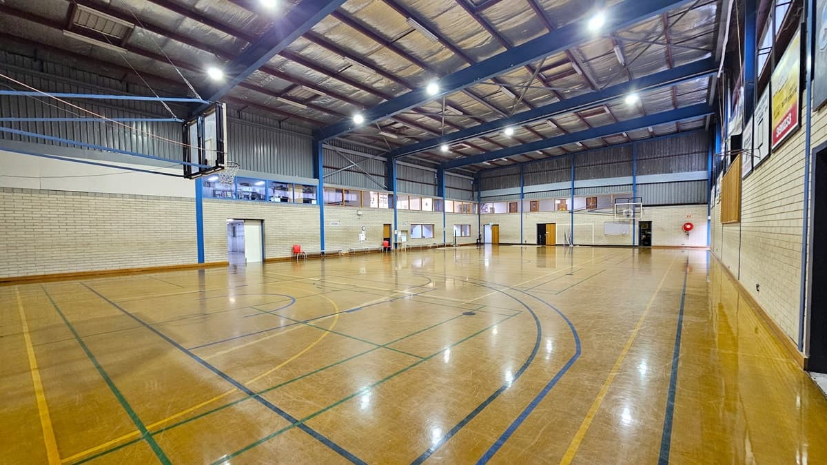 Court 2 features a sturdy concrete floor with game markings, showing signs of worn-out paint in some areas. On the left, bench chairs are available for seating. The court is enclosed by a white brick wall with boards and posters on the right.  The ceiling is equipped with exposed steel beams, fluorescent lights, and insulation foams. Basketball boards are placed on both sides.