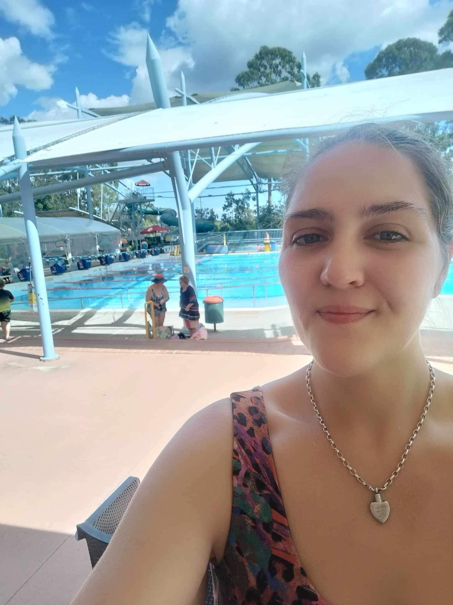 A woman taking a selfie at a poolside area with blue water in the background.
