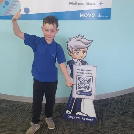 A boy in a blue shirt stands next to a Cérge cartoon cutout with a QR sign.