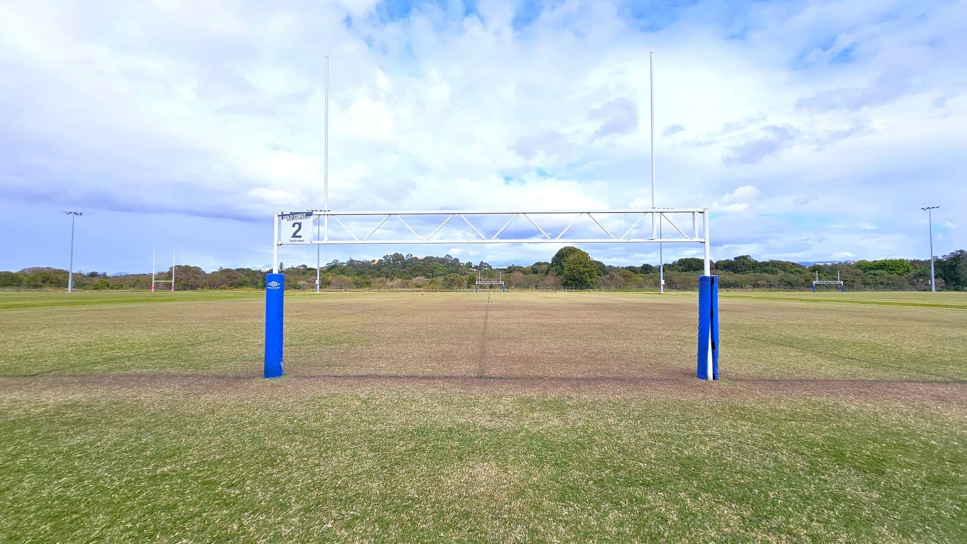 A rugby field with goalposts in the centre, featuring blue padding at the base. The field is mostly grass with some worn patches. In the background, there are more goalposts and a line of trees. The sky is cloudy, and there are floodlights on either side of the field.