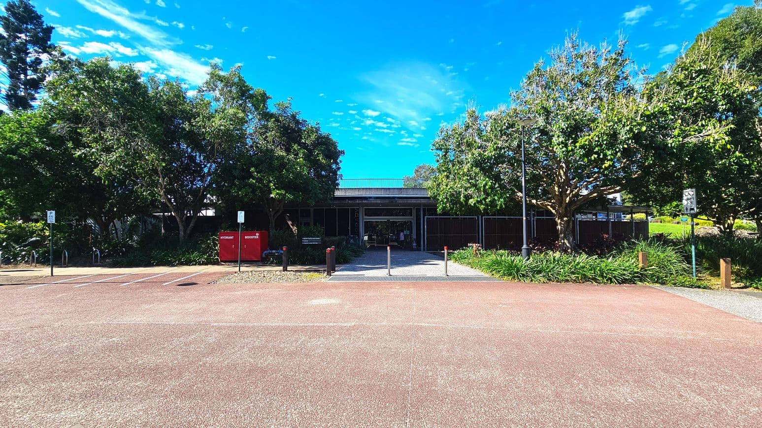 A building entrance is flanked by lush green trees and shrubs. The path leading to the entrance is bordered by bollards and a red hydrant box marked 'HYDRANT BOOSTER' is visible on the left. The building has a flat roof and large glass windows. Parking spaces are marked on the asphalt in the foreground. Signs are posted near the entrance, and a bicycle rack is partially visible on the left.