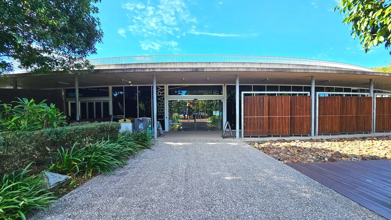 A modern library entrance with a flat, overhanging concrete roof supported by cylindrical columns. The facade features large glass doors flanked by vertical wooden slats. A wide, pebbled pathway leads to the entrance, bordered by lush greenery on the left and a rocky area on the right. A wooden deck is visible on the right side. Signage reading 'LIBRARY' is mounted vertically near the entrance.