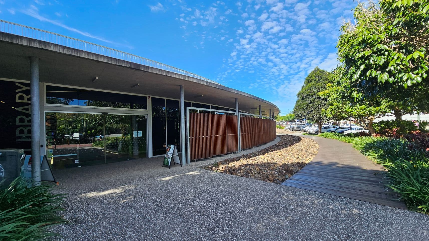 A modern building with a curved facade featuring large glass doors and vertical wooden slats. The entrance is marked by a sign reading 'Library'. A smooth, pebbled pathway leads to the entrance, flanked by a landscaped area with rocks and greenery. To the right, a wooden boardwalk curves through a garden with trees and shrubs. Cars are visible in a parking area beyond the garden.