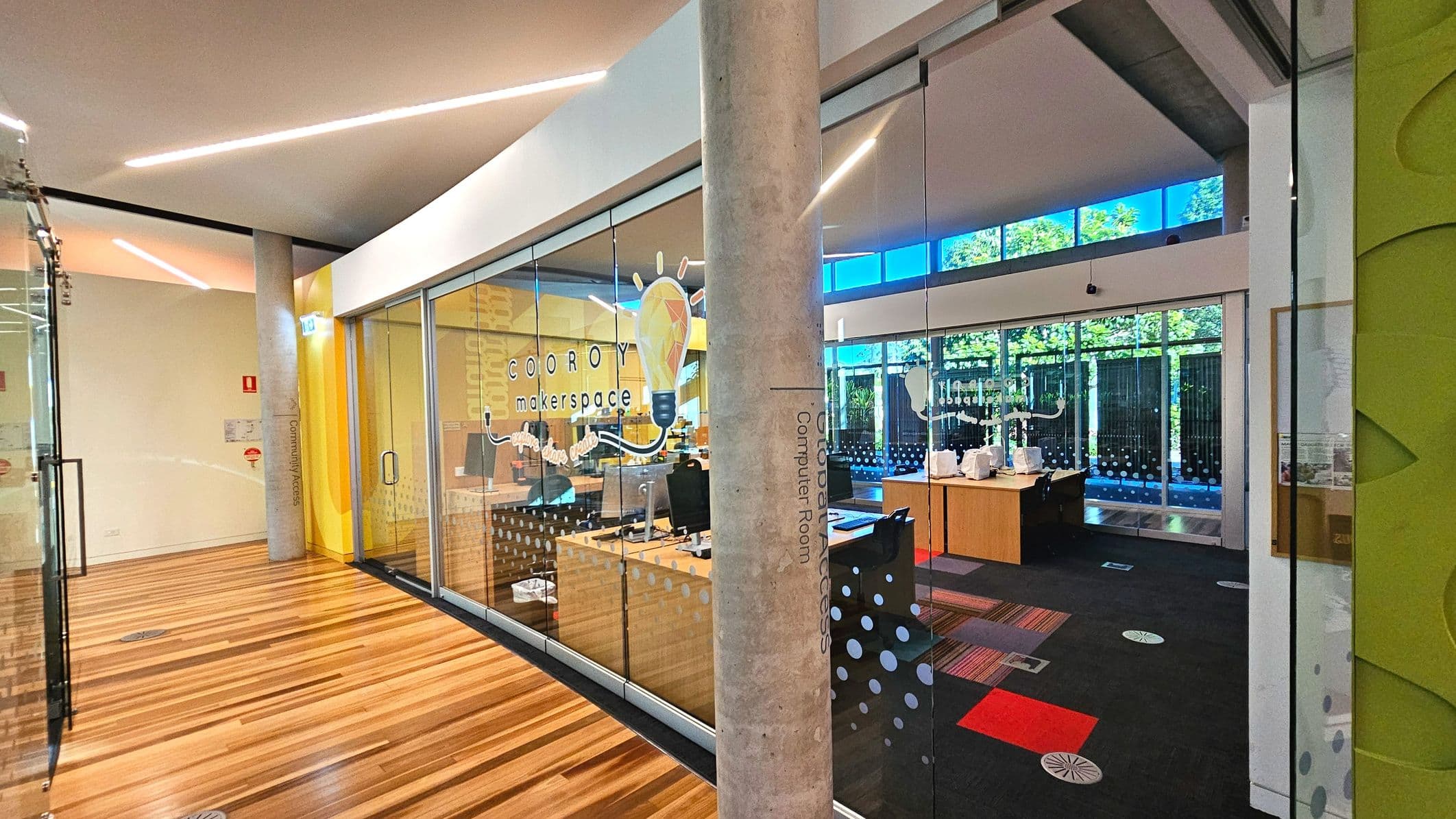 The image shows an interior view of a modern workspace with glass walls labelled 'Cooroy Makerspace'. Inside, there are desks with computers and sewing machines. The floor transitions from polished wood in the foreground to a carpet with red and grey squares inside the room. A concrete pillar stands near the entrance, and large windows at the back let in natural light, revealing greenery outside.