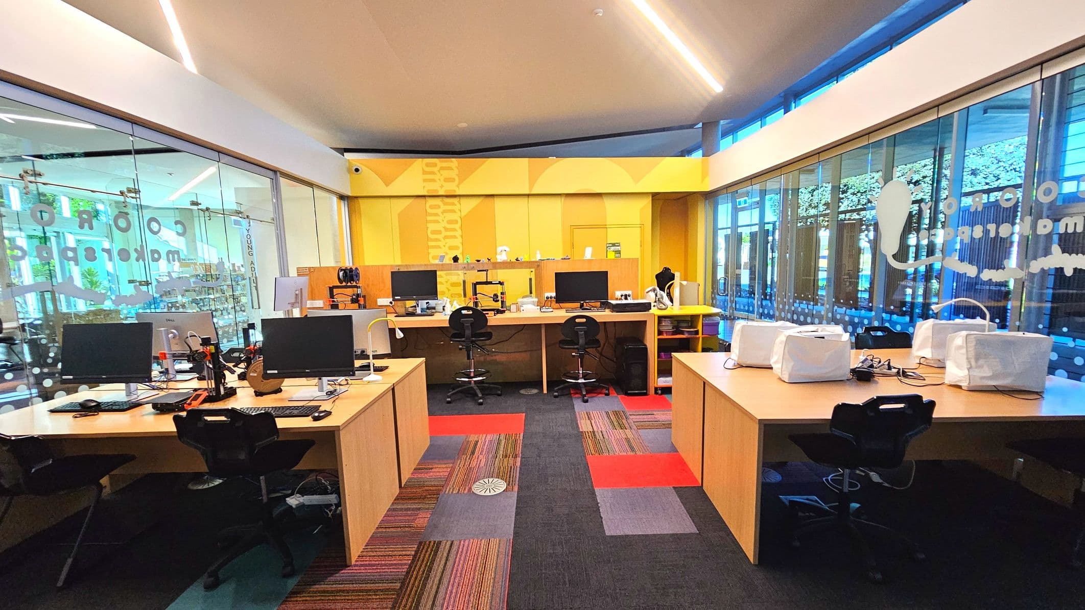 A modern workspace with large glass walls on the left and right, featuring frosted designs and text. Inside, there are several wooden desks arranged in rows, each equipped with computer monitors, keyboards, and various electronic devices. The floor is covered with a colourful patchwork carpet of red, grey, and striped patterns. The back wall is painted yellow with geometric designs, and there are high stools at some desks.