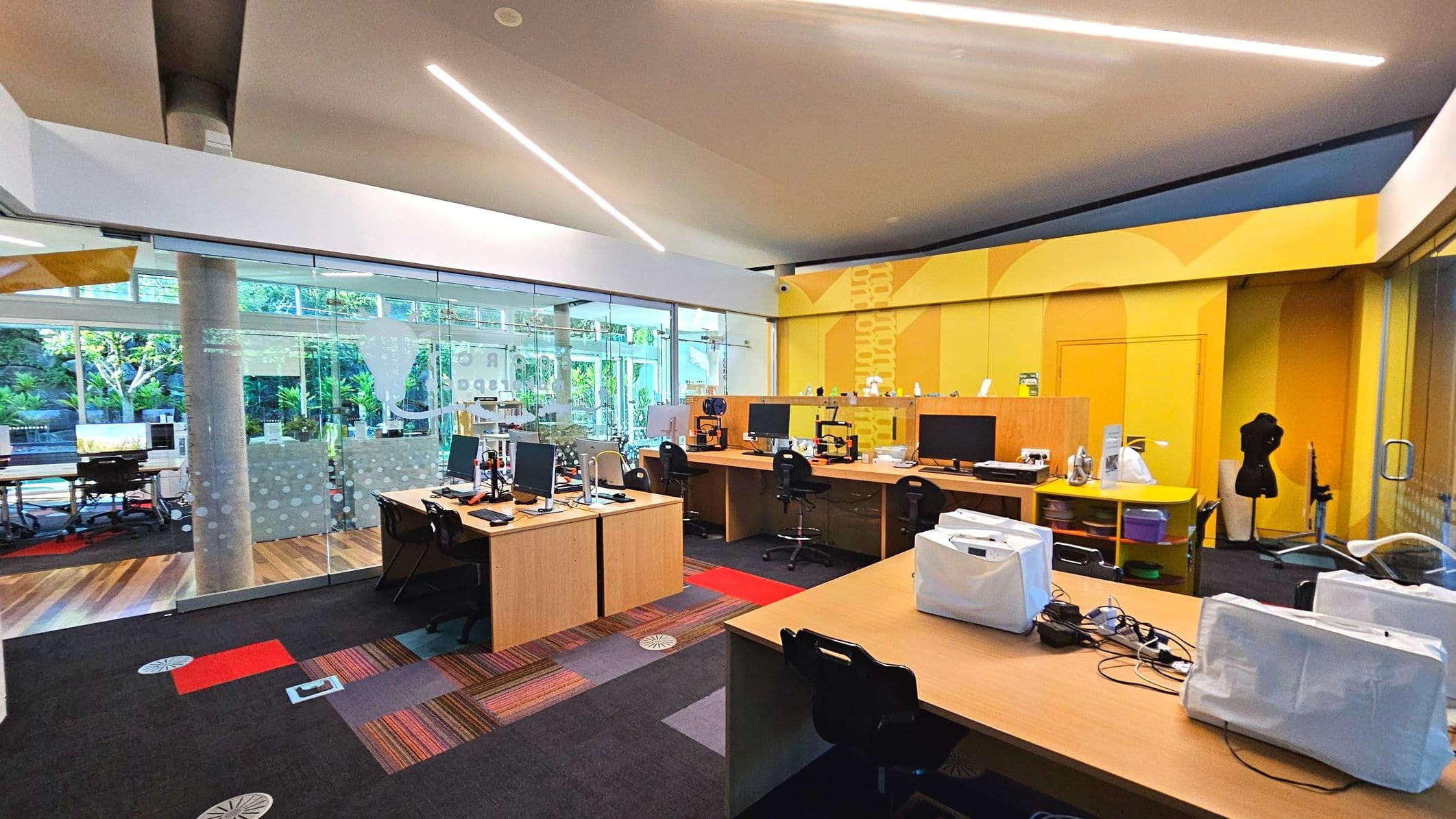A modern office space with a vibrant yellow accent wall and large glass windows revealing greenery outside. The room features several wooden desks equipped with computers and 3D printers. The floor is a mix of dark carpet and colourful patterned sections. A mannequin stands near the right wall. Overhead, sleek linear lights are embedded in the ceiling. The space is open and well-lit, with glass doors providing access.