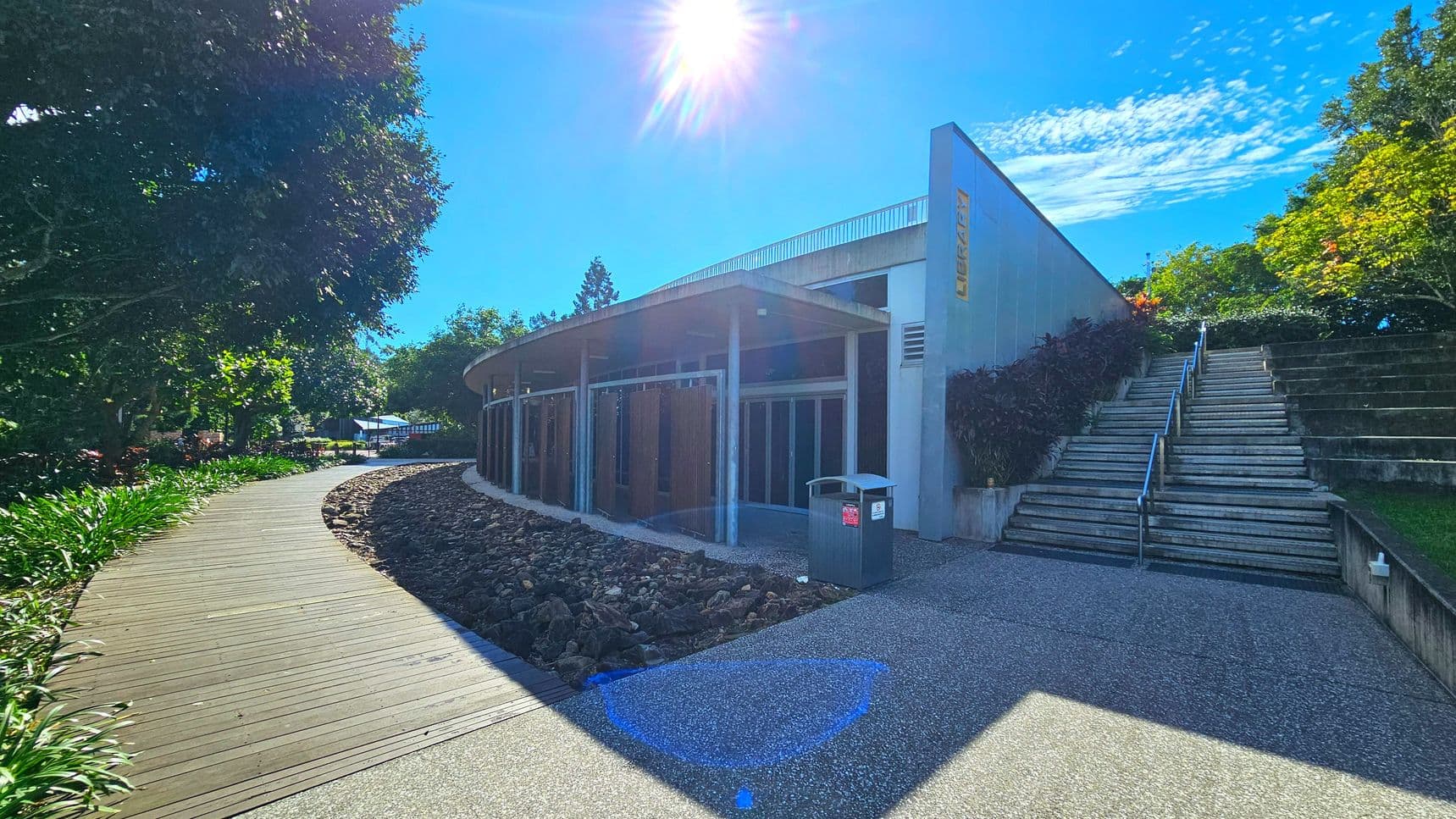 A modern building with a flat roof and large glass doors is situated next to a wide staircase with metal railings. The building is bordered by a wooden boardwalk on the left, which curves through a landscaped area with rocks and greenery. A grey bin is positioned near the entrance. The area feels open, with trees and plants surrounding the structure, and a concrete path leading to the stairs.