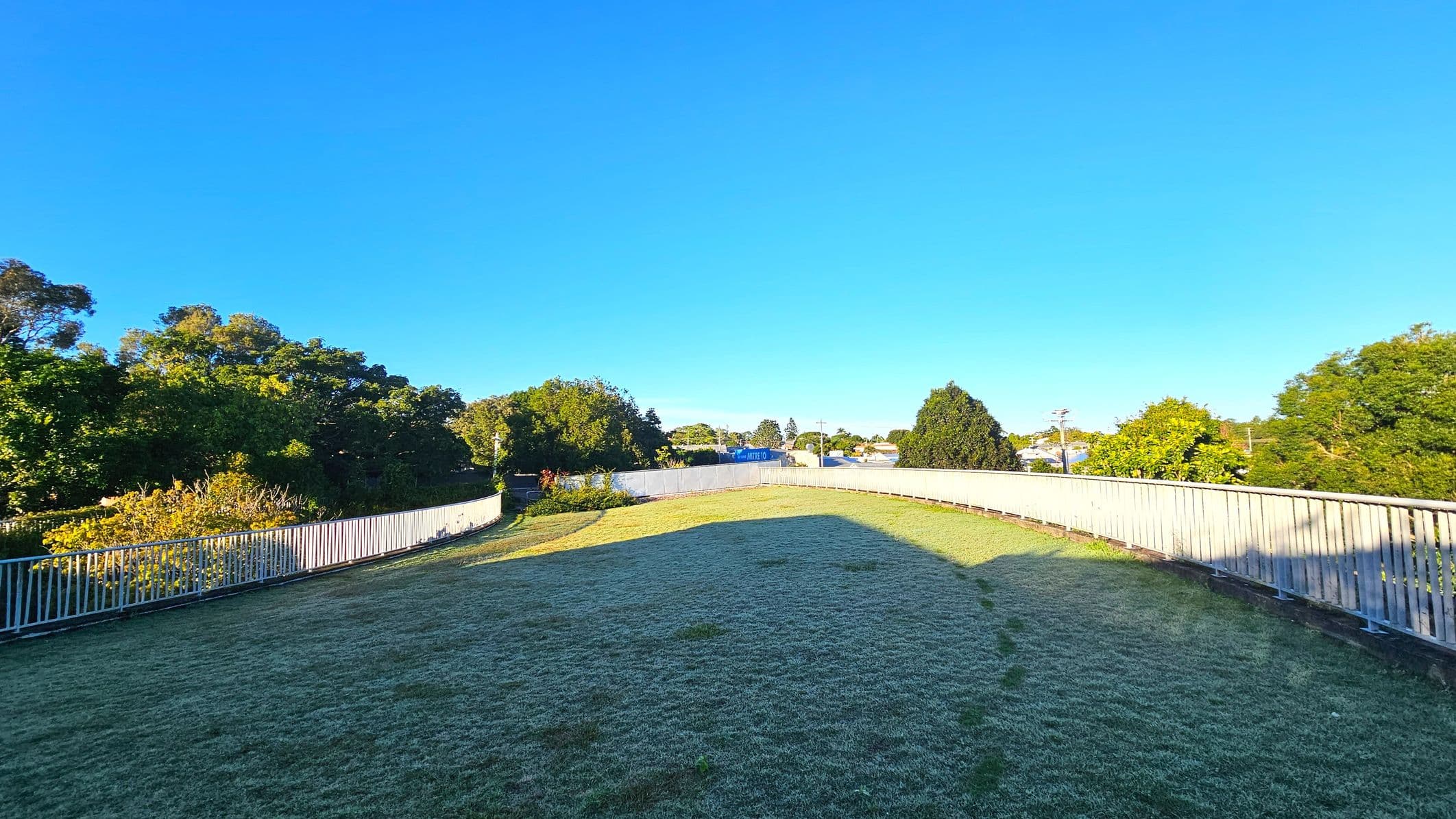 A grassy area with a slight incline is bordered by a white metal railing that curves gently along the perimeter. The grass appears slightly frosted, and there are patches of bare earth. Dense green trees and shrubs surround the area, providing a natural boundary. In the distance, a blue sign is partially visible among the foliage. The space feels open with no visible structures nearby.