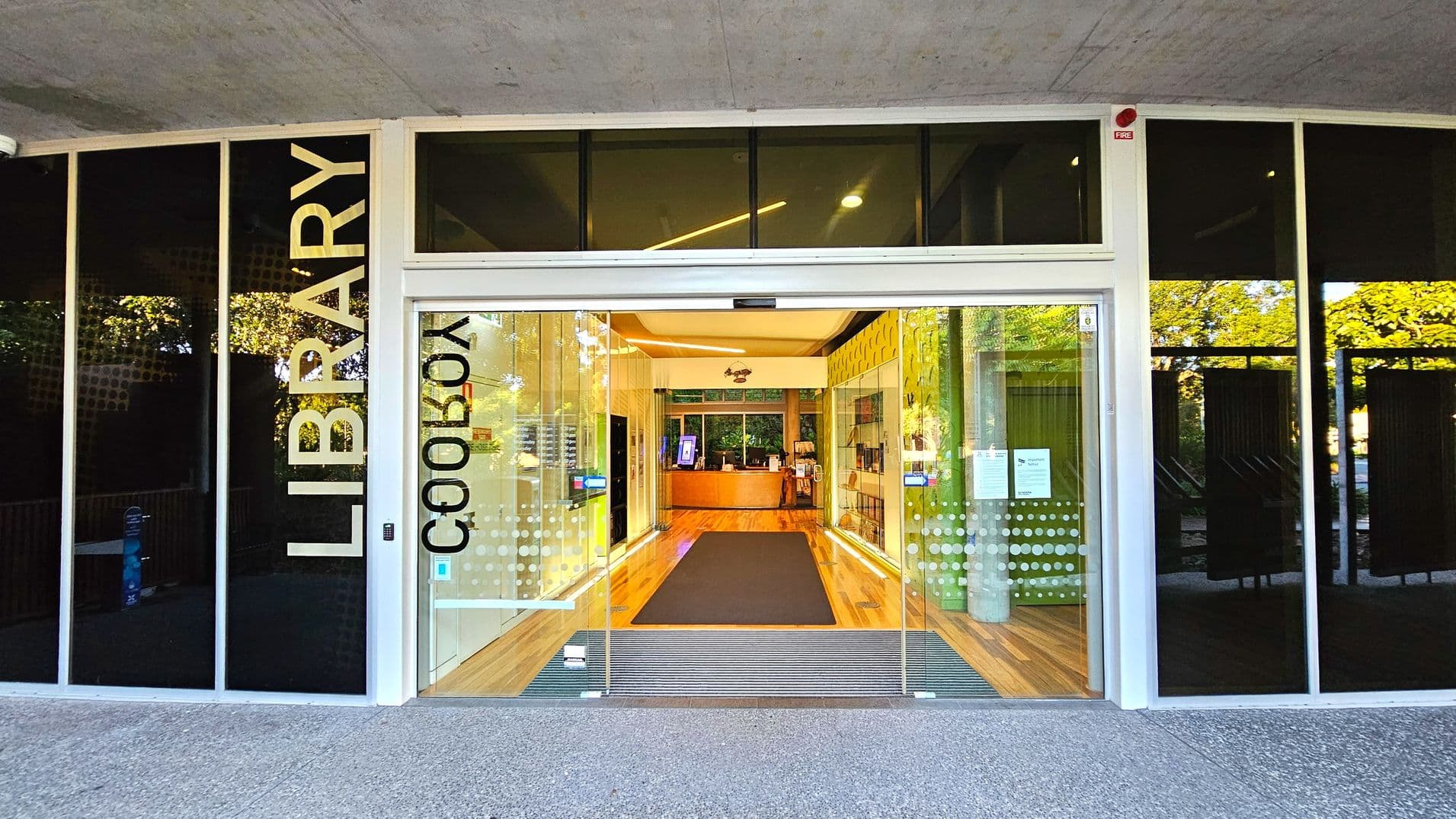 The entrance to a library features large glass doors with the word 'LIBRARY' vertically displayed on the left. The doors are flanked by dark glass panels. Inside, a wooden floor leads to a reception desk with a digital kiosk on the left. The ceiling is light-coloured with recessed lighting. A grey mat is placed at the entrance, and a small step transitions from the outdoor concrete to the indoor wood flooring.