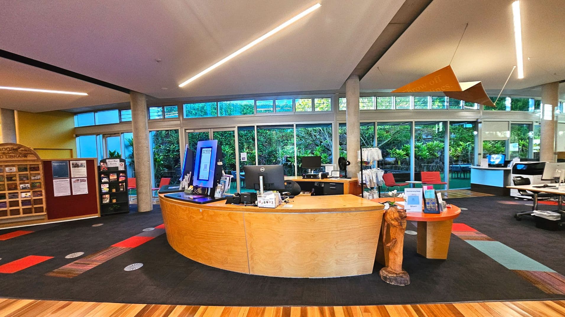 A modern library interior featuring a curved wooden reception desk with computer monitors and informational brochures. Behind the desk, large glass windows reveal lush greenery outside. The floor is a mix of wooden and carpeted surfaces with colourful patterns. Overhead, linear lights and a geometric hanging sculpture are visible. To the left, a wooden display case and a small bookshelf are present.