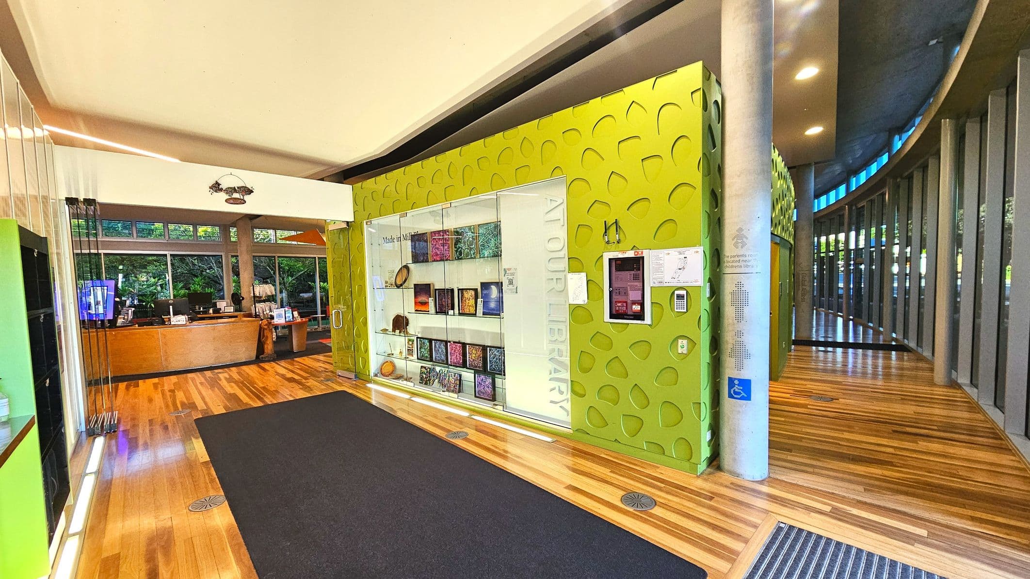 The image shows a modern interior space with a prominent lime green wall featuring a textured pattern and a glass display case showcasing books and artefacts. To the left, a wooden reception desk is visible, backed by large windows with greenery outside. The floor is a mix of light wood and dark carpet. A curved wall with vertical slats and a series of ceiling lights adds to the contemporary design.