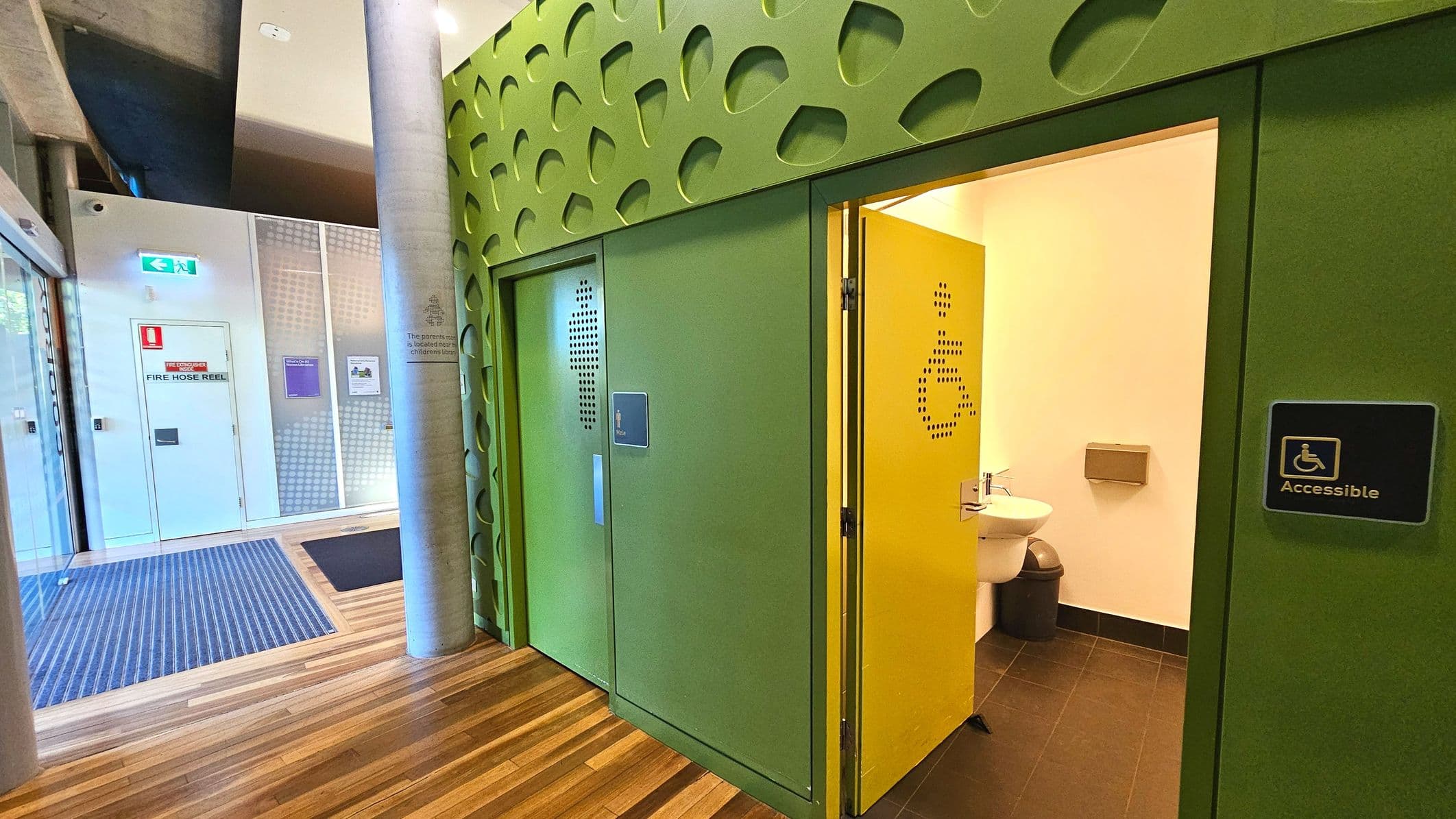 A modern interior space featuring an accessible toilet with a green and yellow colour scheme. The green door has a perforated pattern and an 'Accessible' sign. The yellow door is open, revealing a sink and bin inside. The floor transitions from wood to dark tiles. Nearby, a fire hose reel is mounted on a white wall with a blue exit sign above. A large cylindrical column stands beside the toilet entrance.