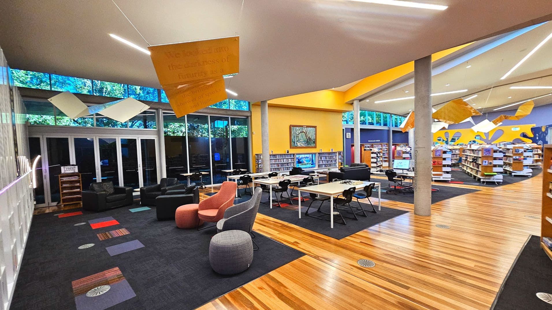 A modern library interior with a mix of seating and shelving. The space features a combination of dark carpet and light wooden flooring. There are several tables with chairs, comfortable armchairs, and bookshelves filled with books. Large windows line one wall, allowing natural light in. The ceiling is adorned with abstract hanging decorations, and a prominent yellow wall adds a splash of colour. A pillar stands centrally, supporting the high ceiling.