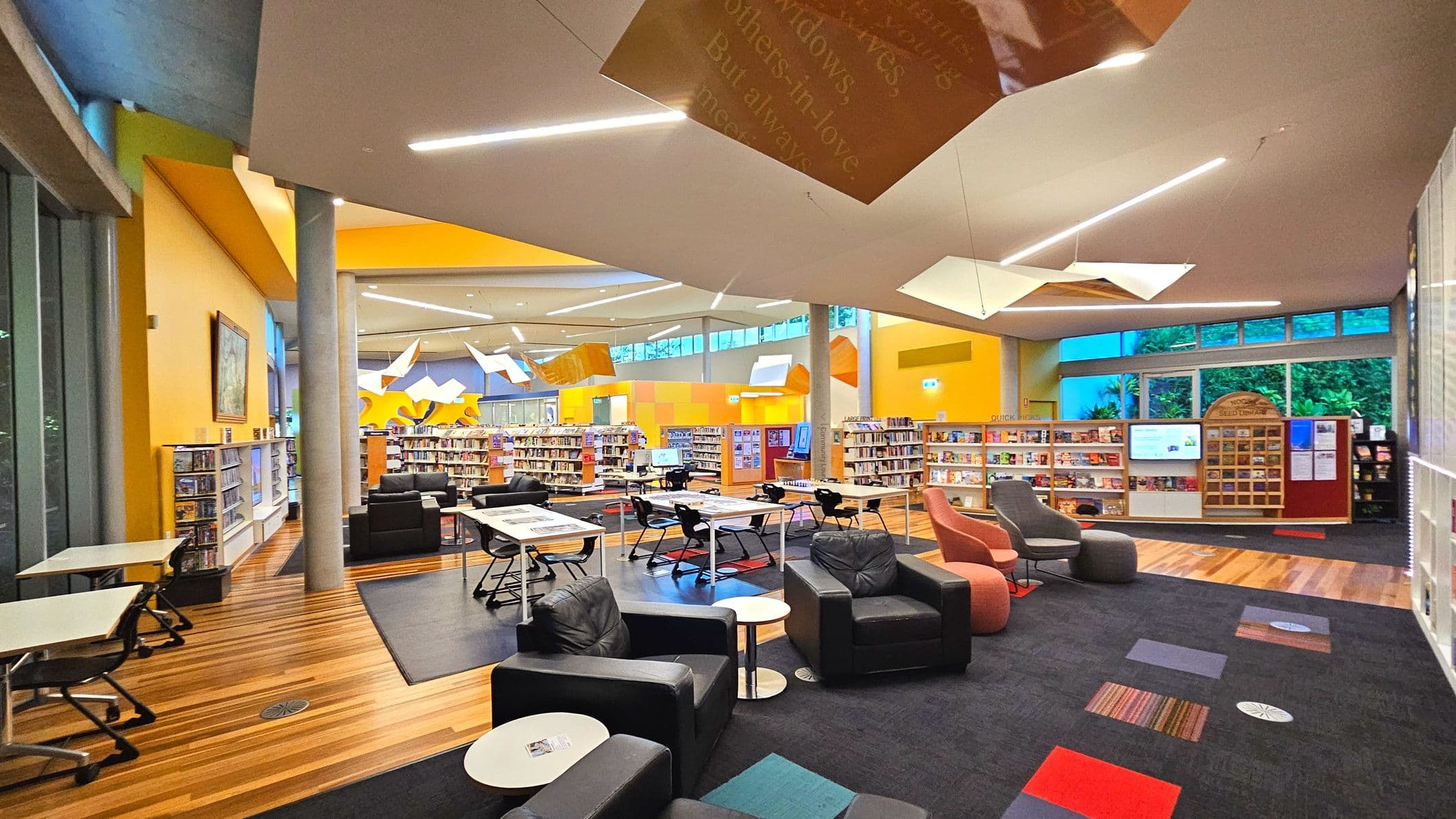 A modern library interior with vibrant yellow and orange walls, featuring a mix of wooden and carpeted flooring. The space is open with high ceilings and geometric light fixtures. Bookshelves line the walls and are arranged in the centre, surrounded by black leather armchairs and tables with chairs. Large windows provide natural light, and a digital screen is mounted on the wall. A column supports the ceiling near the centre.
