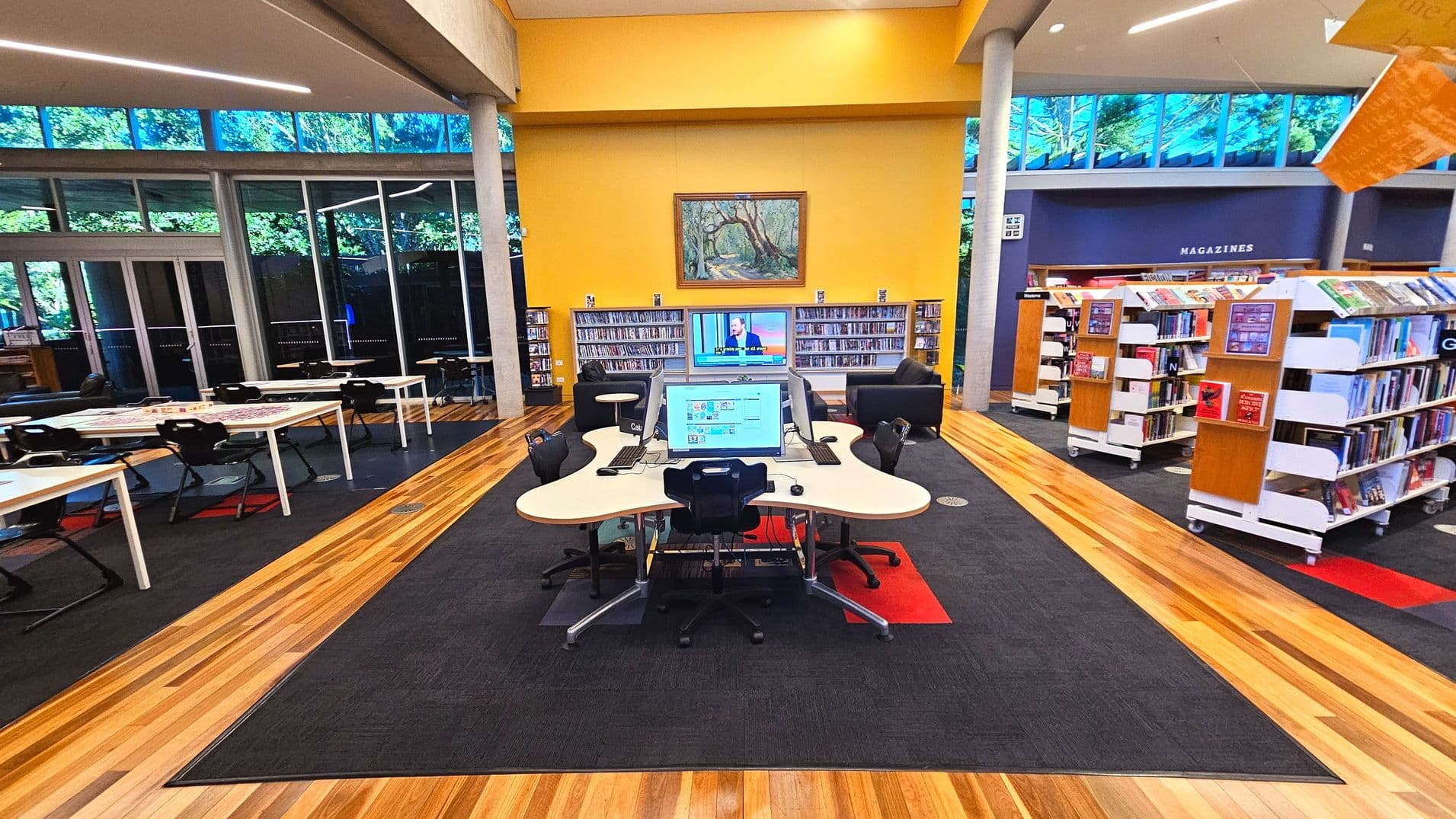 A library interior with a central computer station featuring multiple monitors on a uniquely shaped desk. Surrounding the station are bookshelves filled with books, labelled with letters and a 'MAGAZINES' sign on the wall. The floor is a mix of wooden planks and dark carpet. Large windows with visible greenery outside line the left side, and a yellow wall with a framed painting is at the back.