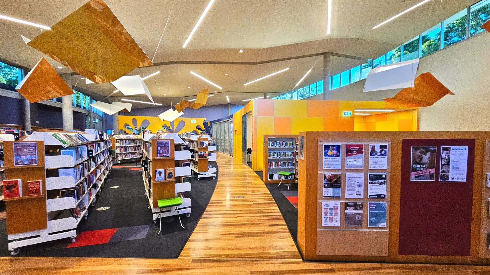 A modern library interior with rows of white and wooden bookshelves filled with books, arranged on a dark carpeted floor. The ceiling features geometric hanging decorations and linear lighting. A prominent yellow and orange wall encloses a section on the right. Large windows near the ceiling allow natural light. Notice boards with flyers are visible on the right. The floor transitions from carpet to light wood.