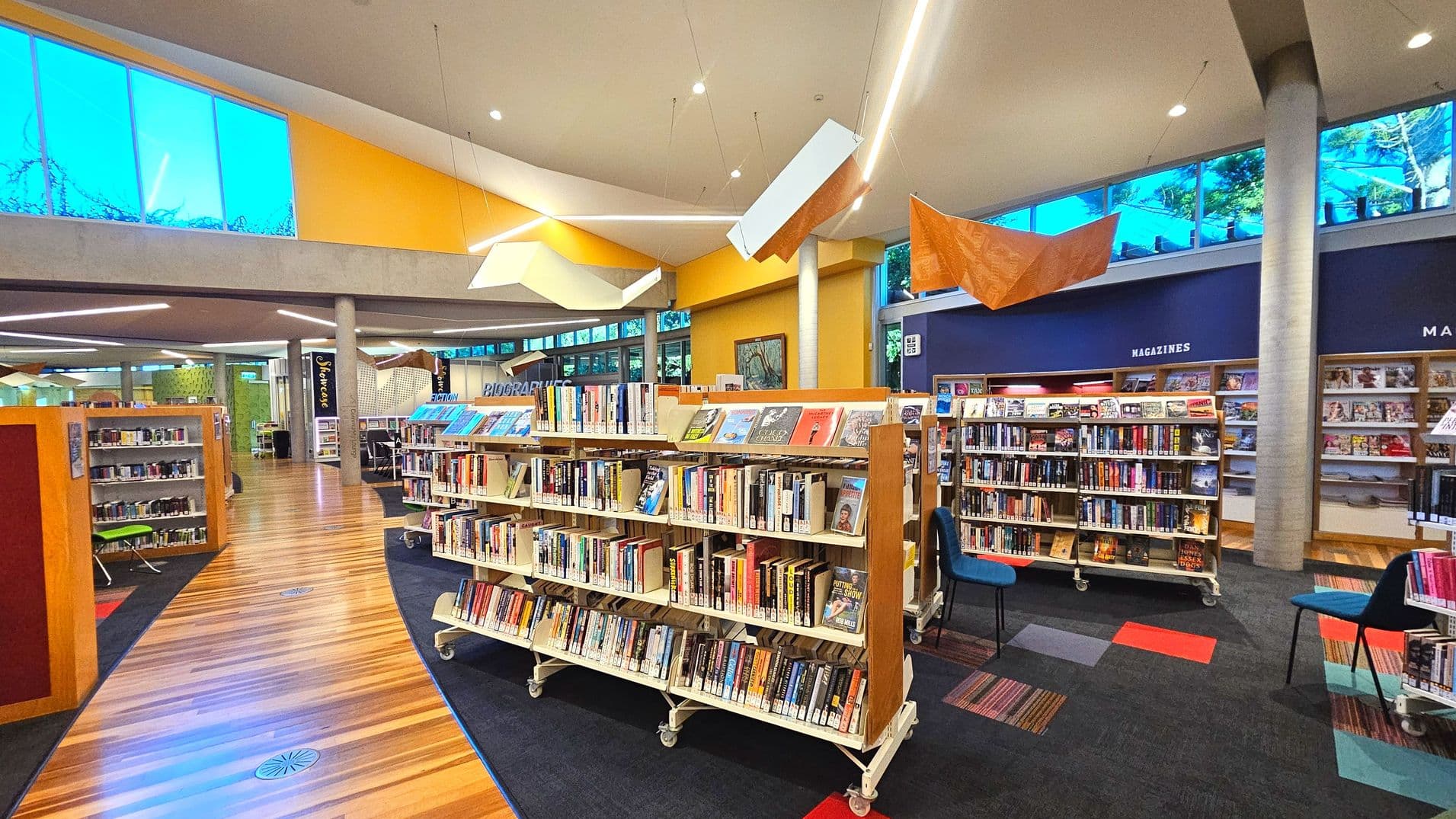 A modern library interior with wooden bookshelves filled with books on a polished wooden floor. The ceiling is high with geometric light fixtures and large windows allowing natural light. The walls are painted in warm yellow and blue tones. There are labelled sections like 'Magazines' and 'Biographies'. Chairs are scattered around, and the space feels open with a mix of carpet and wood flooring.