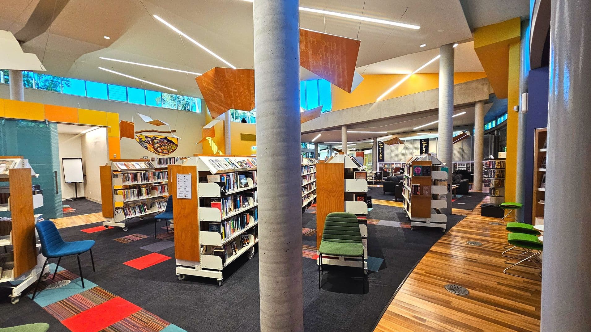 A modern library interior with tall concrete columns and a high ceiling featuring angular, suspended orange panels. Bookshelves are arranged in rows on a dark carpet with red and striped accents. The floor transitions to a wooden surface near green chairs. Large windows near the ceiling allow natural light. A whiteboard is visible in the background, and seating areas are scattered throughout.