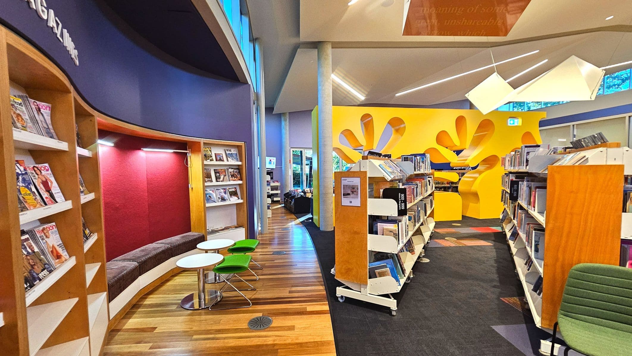 A modern library interior featuring a curved magazine display with a purple backdrop and a red alcove seating area. Adjacent are small round tables with green chairs. The floor transitions from wood to carpet. Bookshelves line the right side, leading to a vibrant yellow wall with abstract cutouts. Large windows and geometric ceiling lights enhance the space's openness.