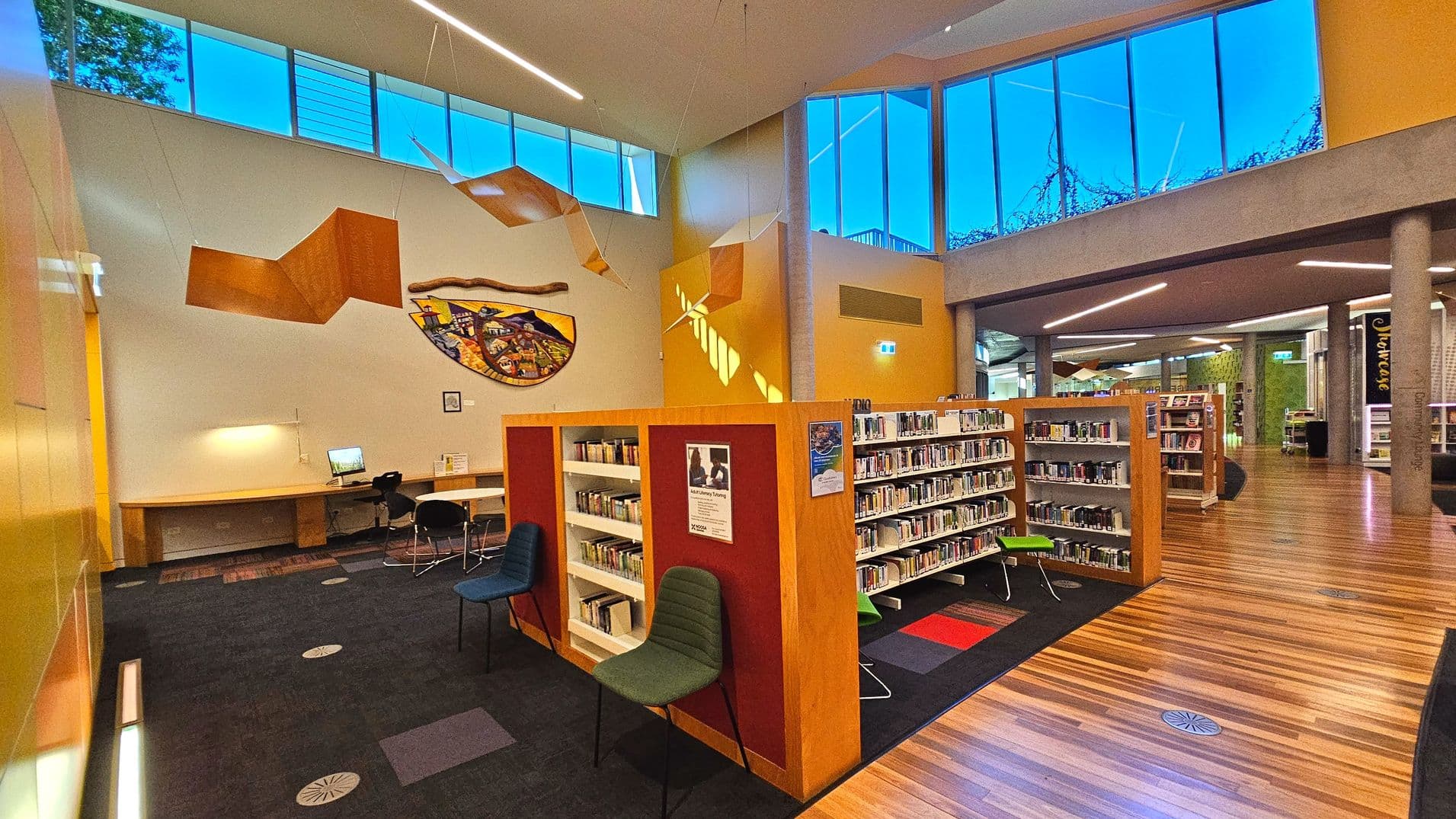 The image shows a modern library interior with wooden bookshelves filled with books, arranged in a semi-open layout. The floor transitions from dark carpet to polished wood. A large, colourful mural adorns the wall above a computer workstation with chairs. High windows allow natural light, and geometric ceiling decorations add a contemporary touch. The space feels open with visible columns and a mix of materials.