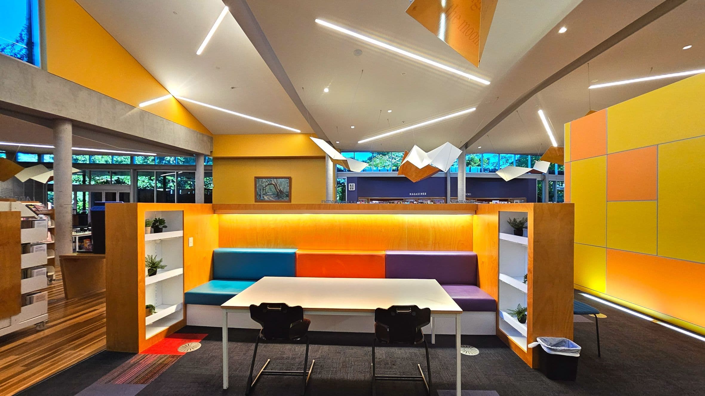 A modern library interior featuring a vibrant seating area with a white table and two black chairs. The seating booth has colourful cushions in blue, orange, and purple, flanked by white shelves with small plants. The walls are adorned with yellow and orange panels. Overhead, angular light fixtures and geometric ceiling designs add a contemporary touch. Large windows provide natural light, and bookshelves are visible in the background.