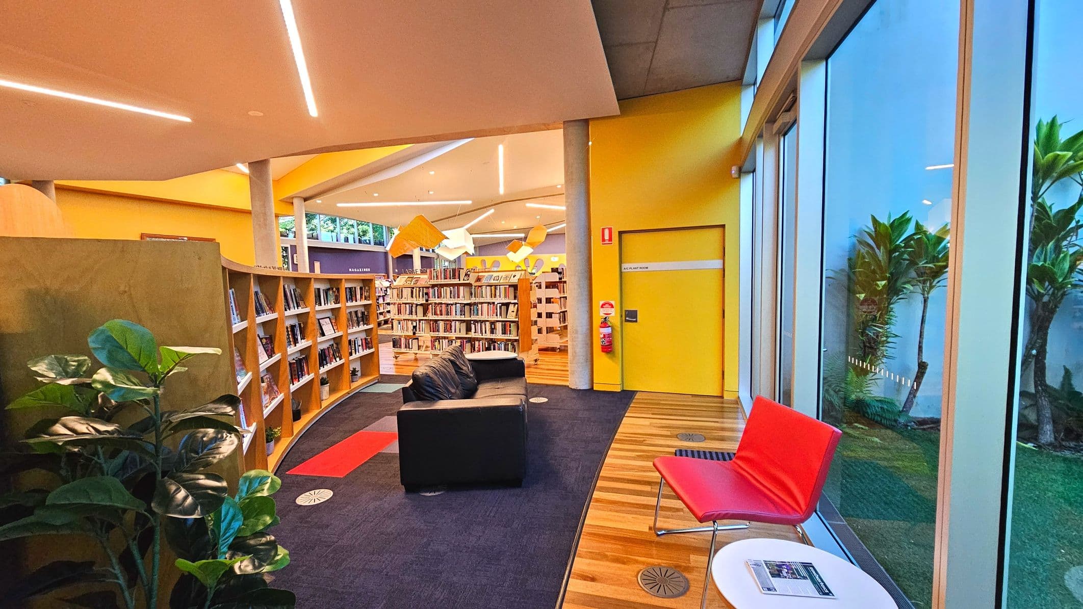 A modern library interior with a vibrant yellow wall and a door marked 'Plant Room'. Bookshelves filled with books curve along the left side, leading to a central area with more shelves. A black sofa and a red chair sit on a carpeted area, transitioning to wooden flooring near large glass windows. Outside, greenery is visible. Overhead, angular ceiling panels with embedded lights add a contemporary touch.