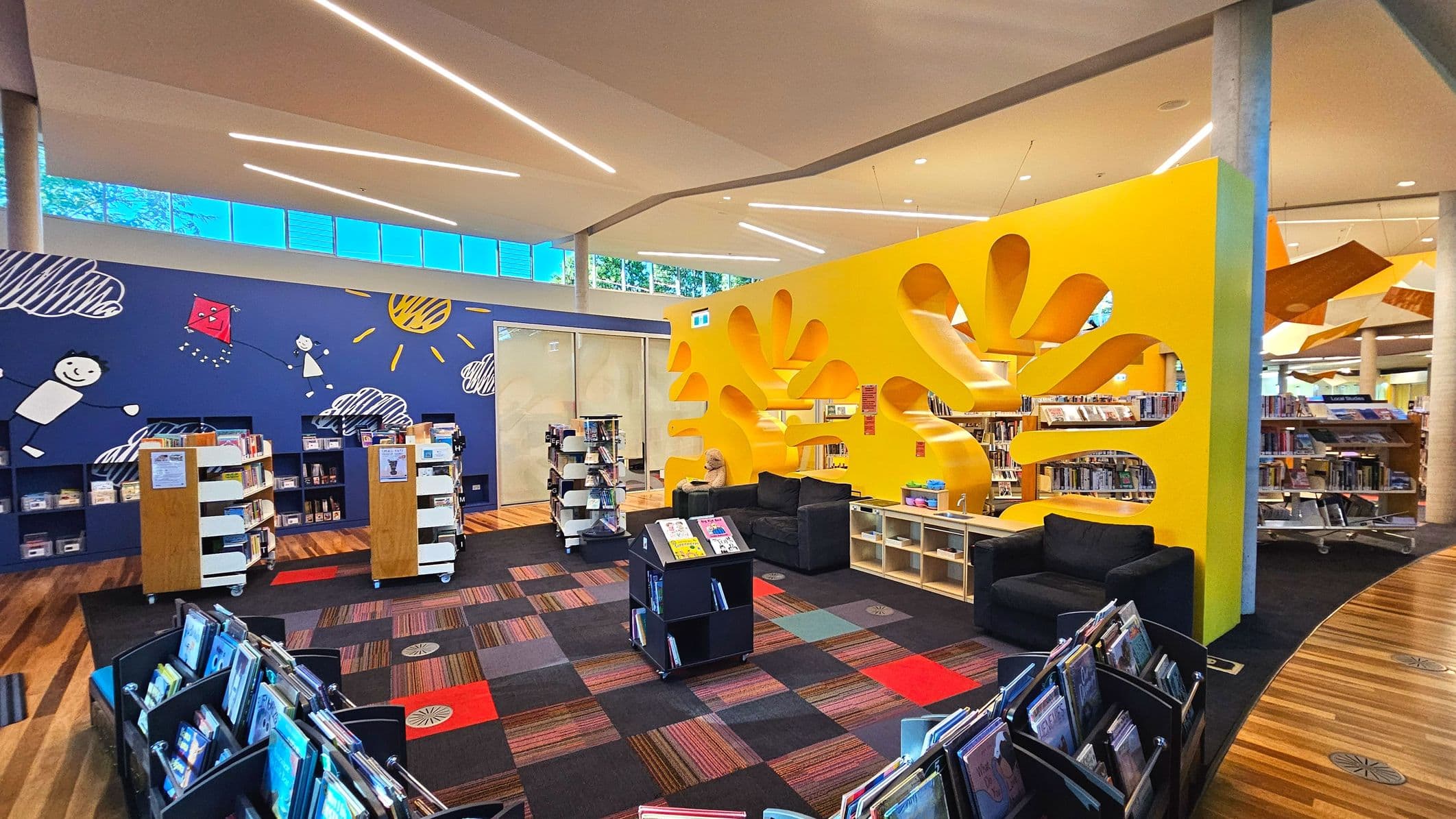 A vibrant library space featuring a large yellow partition with abstract cut-outs, creating a playful atmosphere. The floor is a mix of wooden planks and patterned carpet tiles. Bookshelves are arranged neatly, with a blue wall adorned with cheerful illustrations of children, clouds, and a sun. Comfortable black sofas are positioned for seating. The ceiling has modern linear lighting, and large windows allow natural light in.
