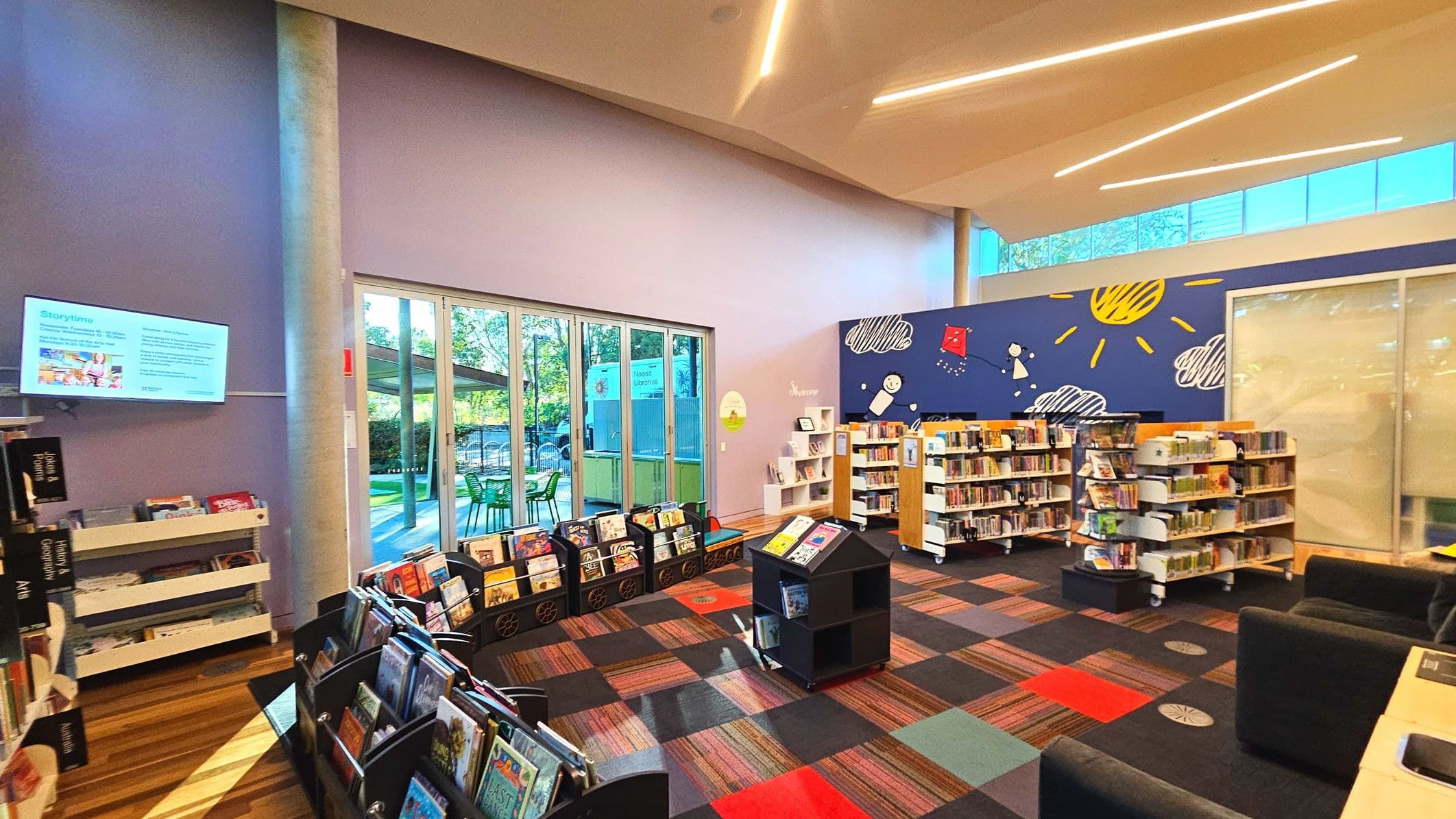 A vibrant library space with colourful carpet tiles and a playful mural featuring a sun and clouds on a blue wall. Bookshelves filled with children's books are arranged neatly. A digital screen displays information on a purple wall. Large glass doors lead to an outdoor area with green chairs and tables. The ceiling has modern, angular lighting, and the space feels open with ample natural light.