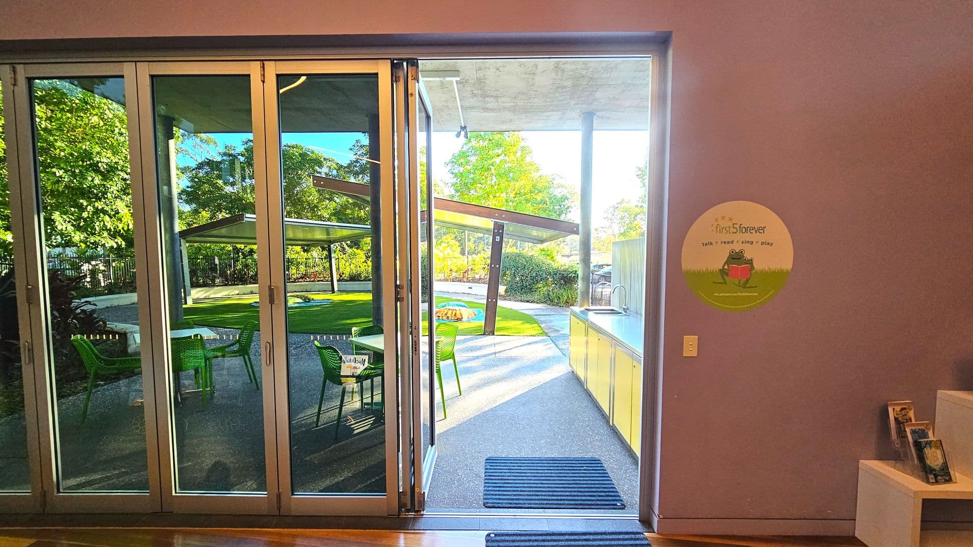 A view through large glass sliding doors leading to an outdoor area with green chairs and tables on a paved surface. Beyond, a grassy area is partially covered by a modern, angular metal canopy supported by poles. Inside, a wall displays a circular sign with a frog illustration. A countertop with cabinets runs along the right side, and a small display shelf holds brochures. The space feels open and connected to the outdoors.