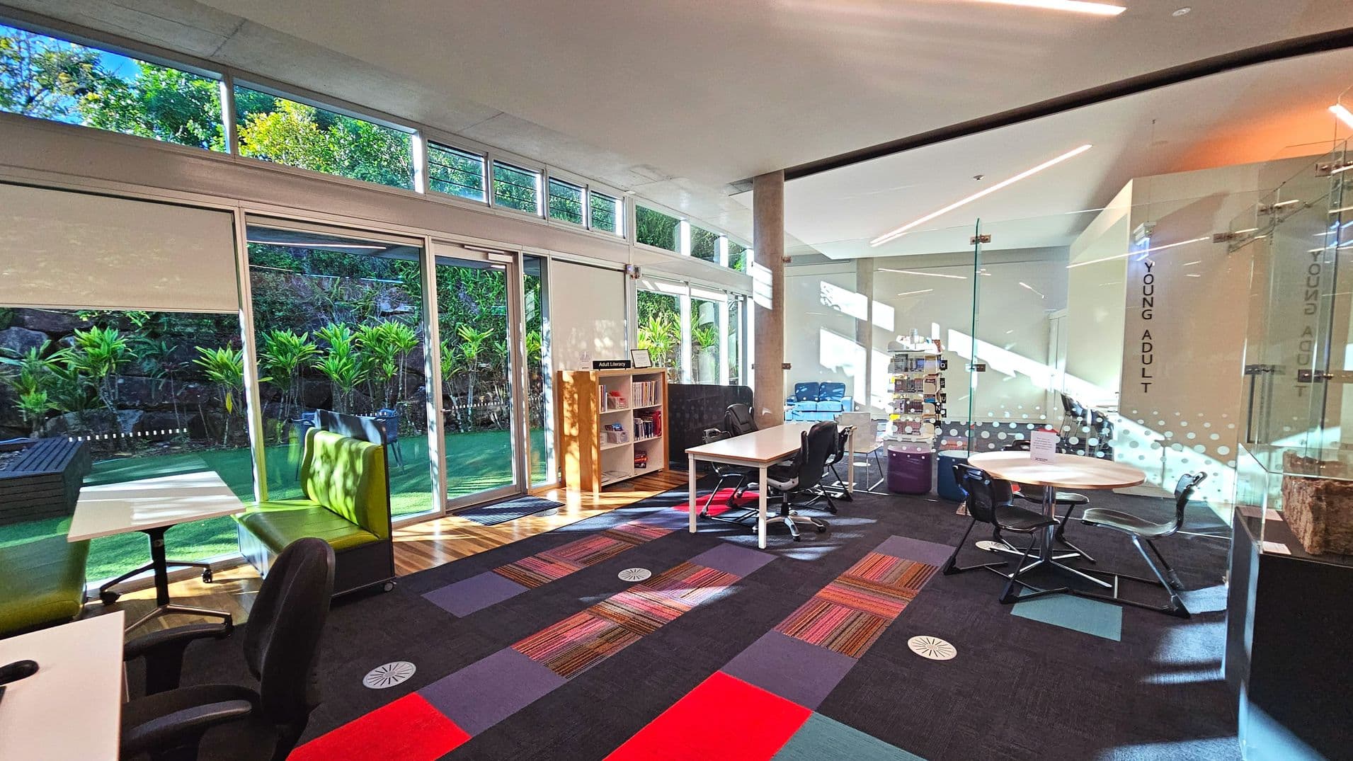 A modern library interior with large glass doors and windows on the left, revealing a lush garden outside. Inside, there are tables with chairs, a green cushioned bench, and a small bookshelf labelled 'Adult Literacy'. The floor features a colourful carpet with geometric patterns. A glass wall on the right has 'Young Adult' signage. The space is well-lit with natural light streaming in.