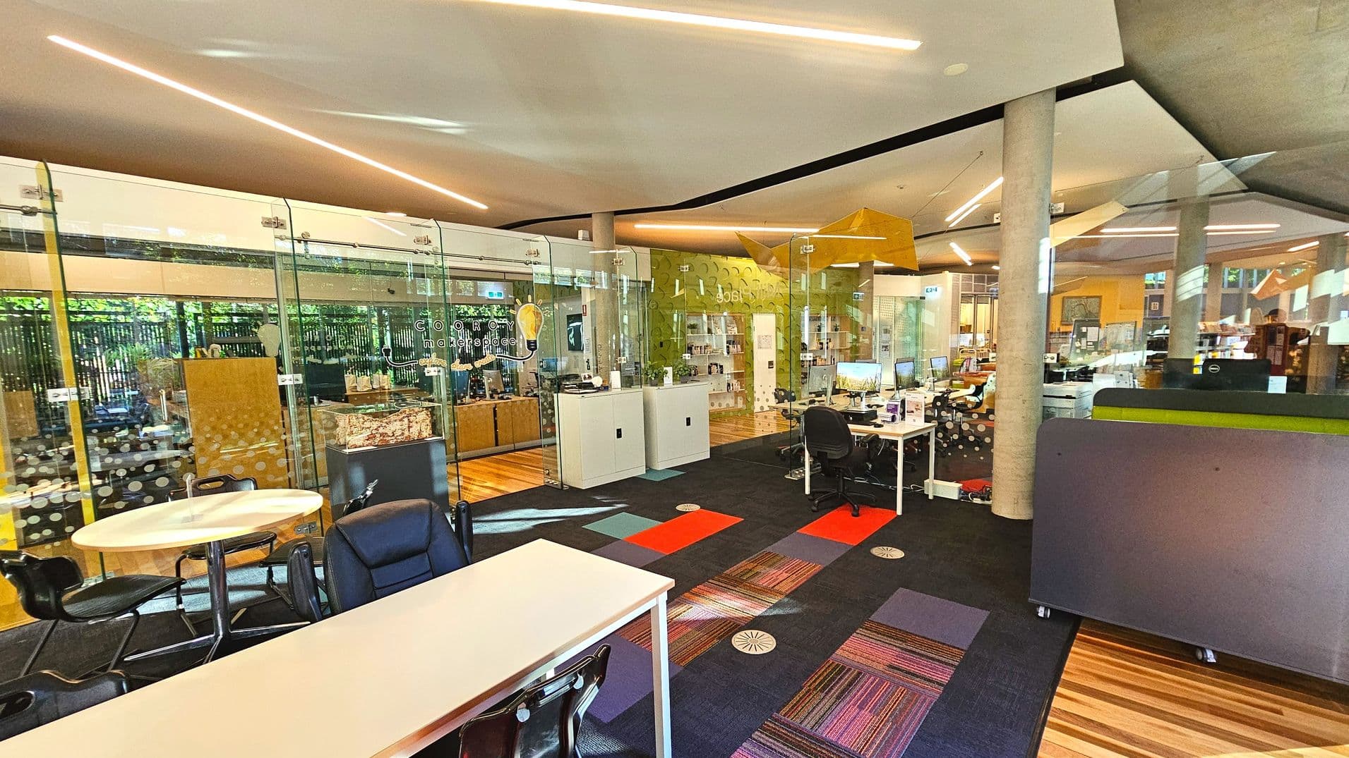 The image shows a modern office space with glass walls and a mix of open and enclosed areas. There are several desks with computers, chairs, and a round table with black chairs in the foreground. The floor features a combination of wooden and carpeted surfaces with colourful patterns. A large pillar supports the ceiling, and there are decorative elements like a yellow geometric structure hanging from above.