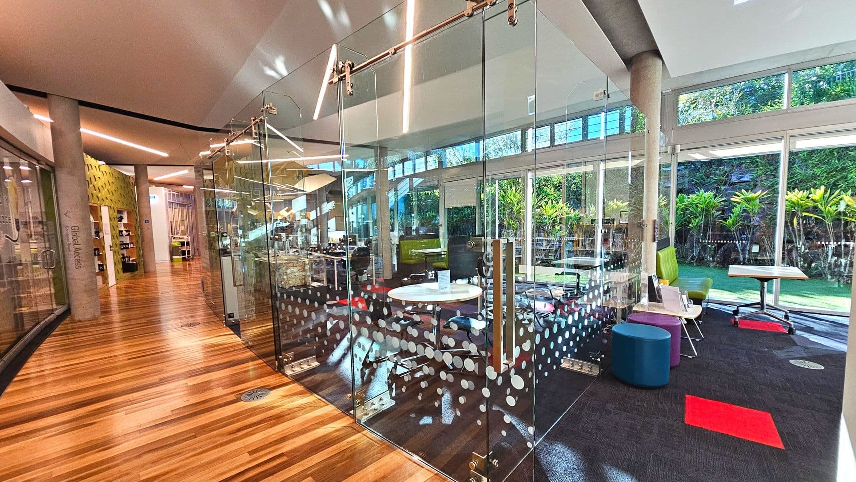 A modern interior space featuring a glass-walled meeting room with sliding doors, adorned with a pattern of white dots. Inside, there's a round white table and office chairs. The room is adjacent to a corridor with polished wooden flooring and concrete pillars. Large windows provide views of greenery outside. The area includes colourful seating and a mix of carpet and wood surfaces, creating a vibrant, open atmosphere.