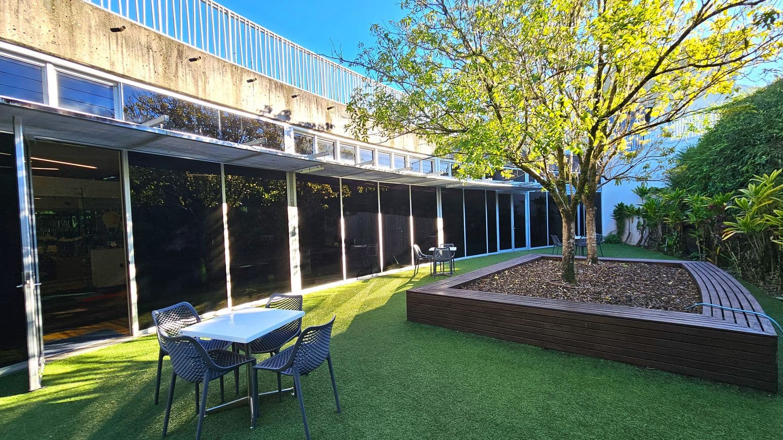 An outdoor courtyard features a large tree planted in a raised wooden planter with a curved bench. The ground is covered with artificial grass. Adjacent to the planter are two sets of tables and chairs. A building with large glass windows and a concrete upper section with a railing runs along one side. The area is bordered by dense greenery, creating a secluded atmosphere.