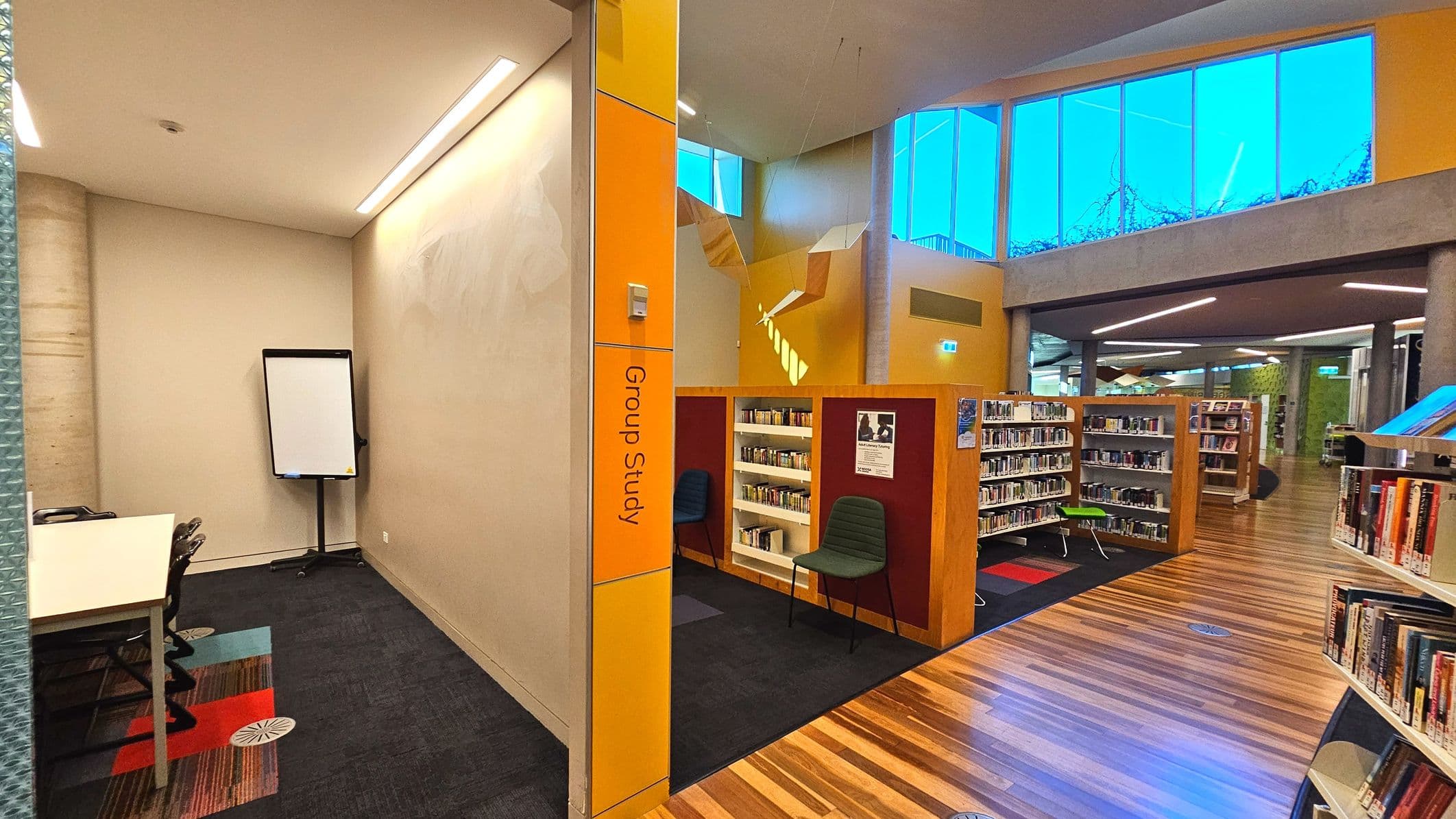 A library interior featuring a 'Group Study' area marked by a yellow sign on a wall. To the left, a small room with a whiteboard and chairs on a carpeted floor with colourful squares. Adjacent are bookshelves with books and a chair. The space opens into a larger area with wooden flooring and more bookshelves. Large windows above allow natural light, and the ceiling is high with modern lighting.