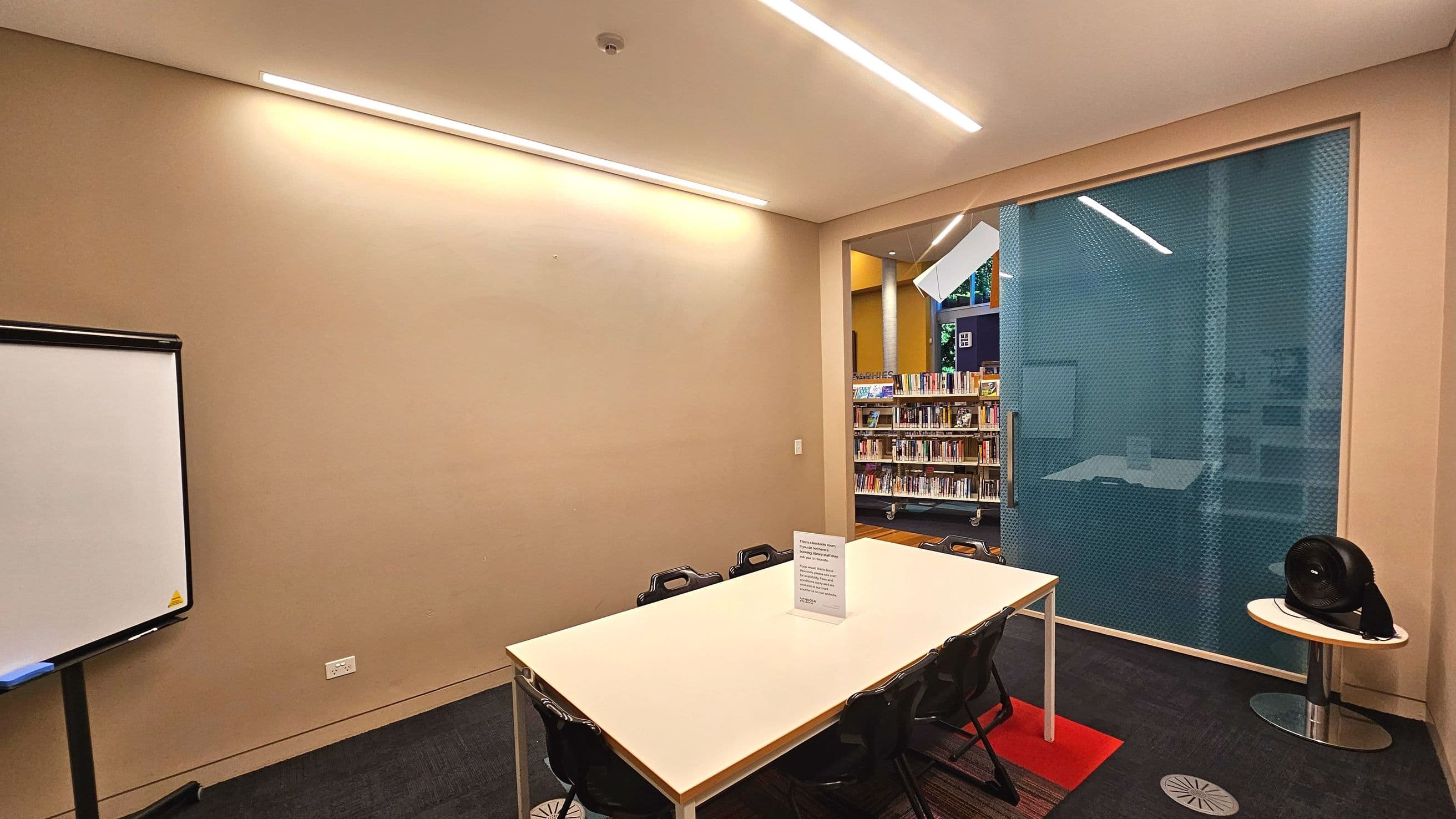 A small meeting room with a rectangular white table surrounded by black chairs. A whiteboard stands to the left against a beige wall. The room is partially enclosed by a large glass panel with a blue tint, through which a library area with bookshelves is visible. The floor is carpeted in dark grey with a red mat under the table. A small round table with a black fan is positioned to the right.