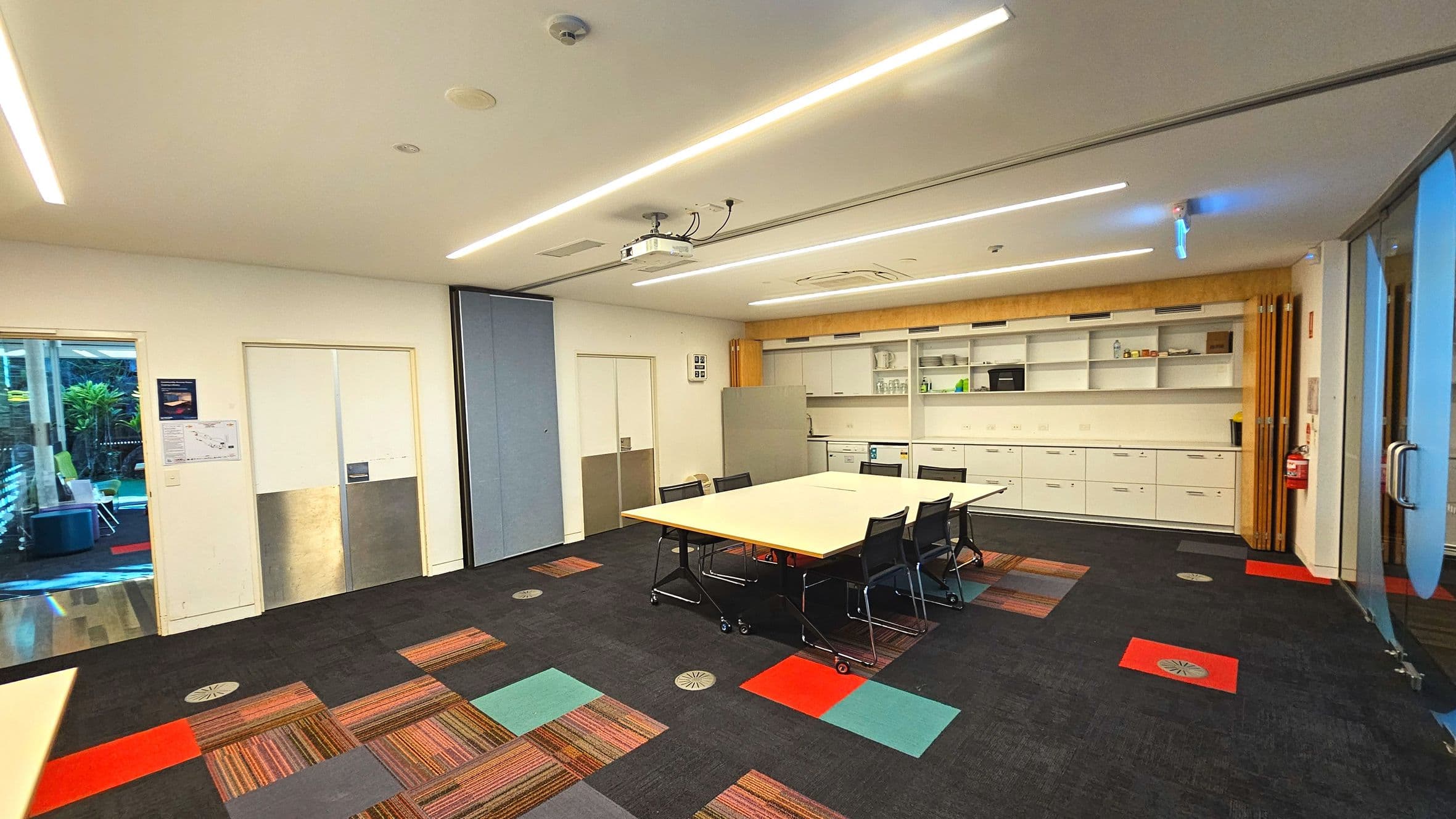 A modern meeting room with a rectangular table surrounded by black chairs on wheels. The floor features a patchwork of colourful carpet tiles in red, blue, and striped patterns. A ceiling-mounted projector is above the table. The back wall has white cabinets and open shelves with various items. A glass door on the right and a sliding door on the left lead to other areas. The room is well-lit with linear ceiling lights.